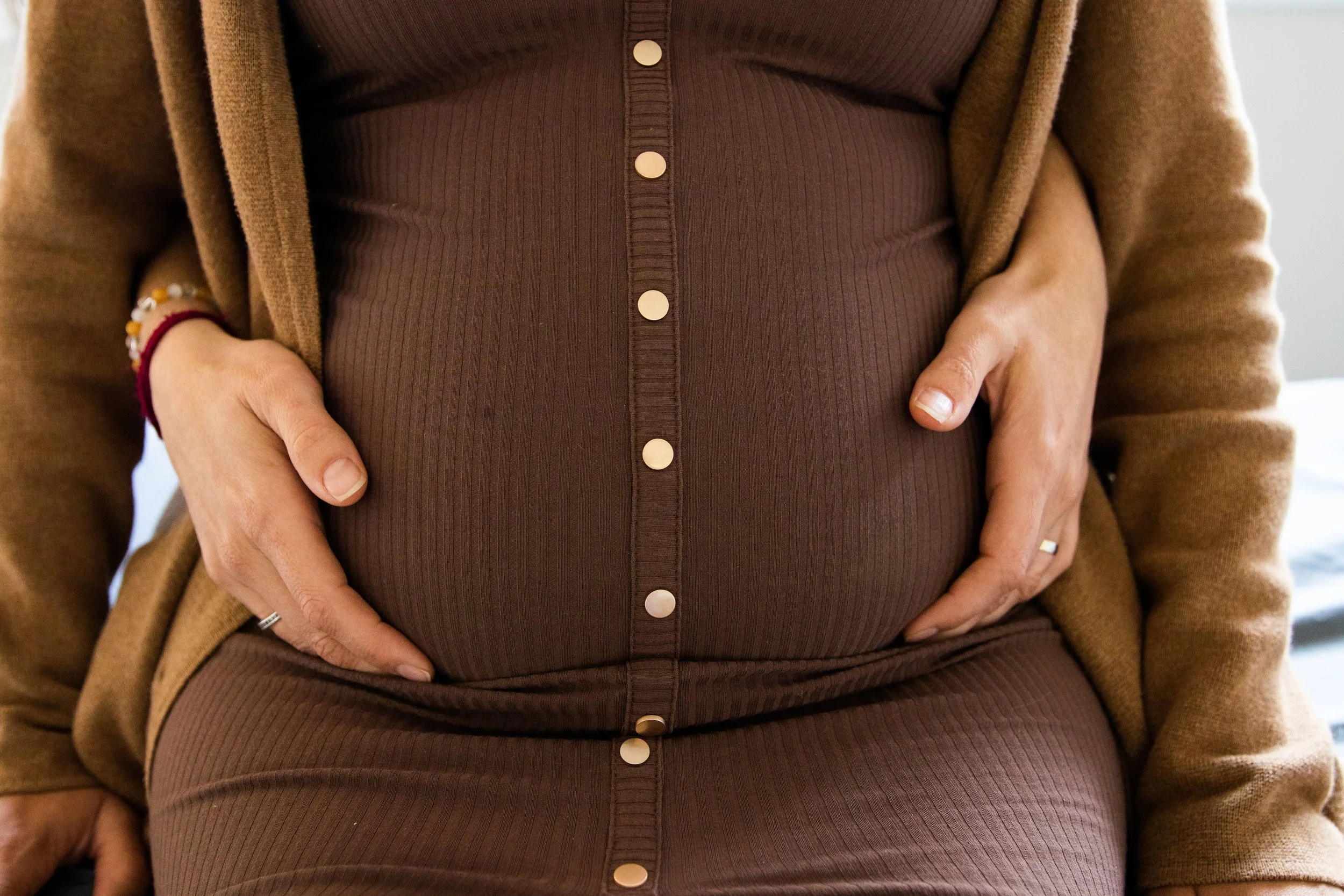 A woman with dark skin, light brown hair, wearing a blue polka dot shirt and beige pants, sitting on a chair with folded arms, smiling and talking to a man with dark skin, blonde dreadlocks, wearing a white shirt and black shorts, sitting on a beige sofa, with a laptop on the wooden shelf in the background.