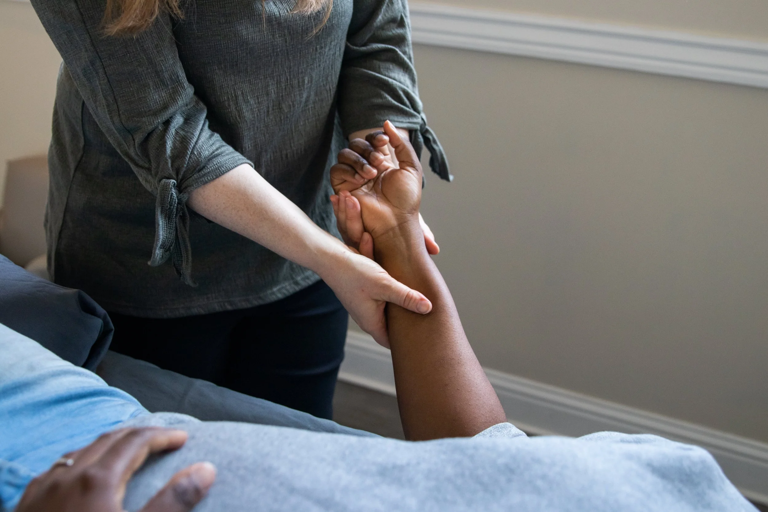A person lying in bed being assisted by another person who is holding their arm and hand, possibly in a medical or caregiving context.