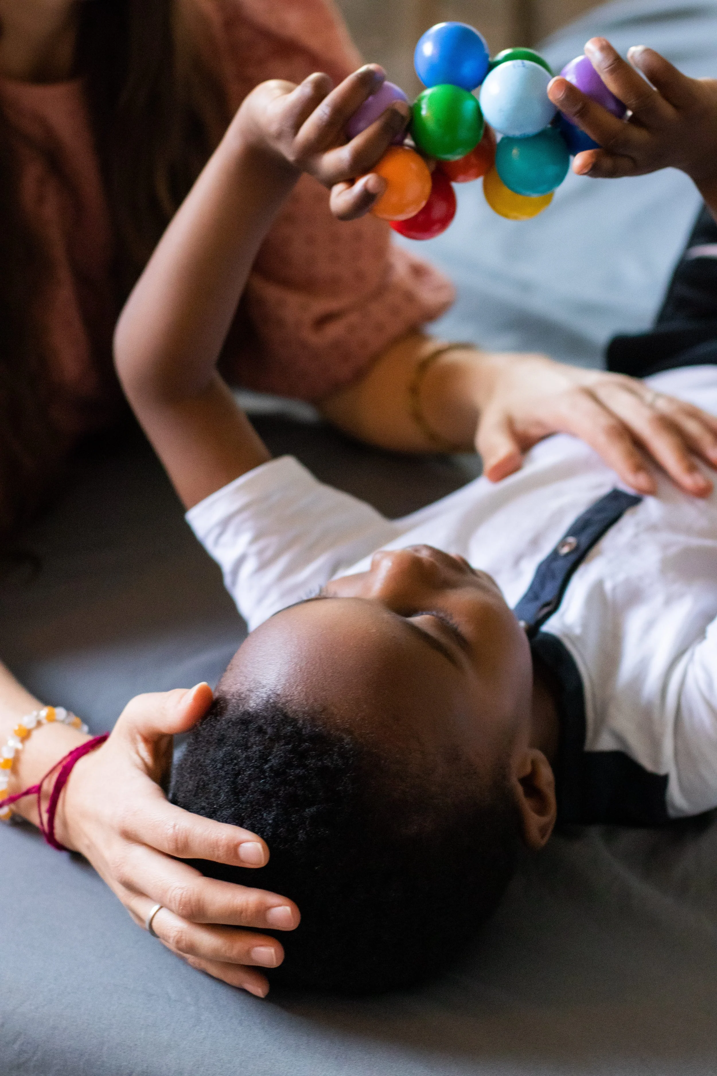 A woman with dark skin, light brown hair, wearing a blue polka dot shirt and beige pants, sitting on a chair with folded arms, smiling and talking to a man with dark skin, blonde dreadlocks, wearing a white shirt and black shorts, sitting on a beige sofa, with a laptop on the wooden shelf in the background.