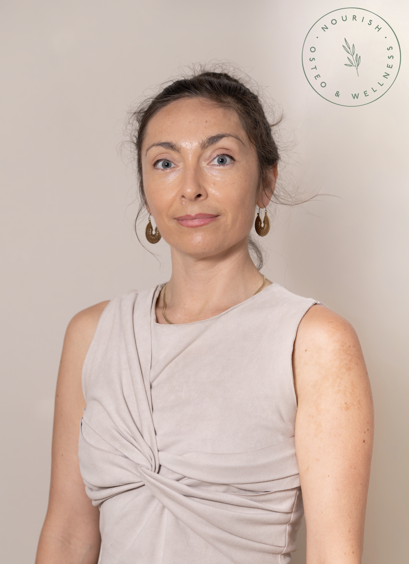 A woman with dark brown hair, wearing a white button-up shirt, gold necklace, and earrings, standing against a brick wall, with her hand on her chest and a gentle smile.