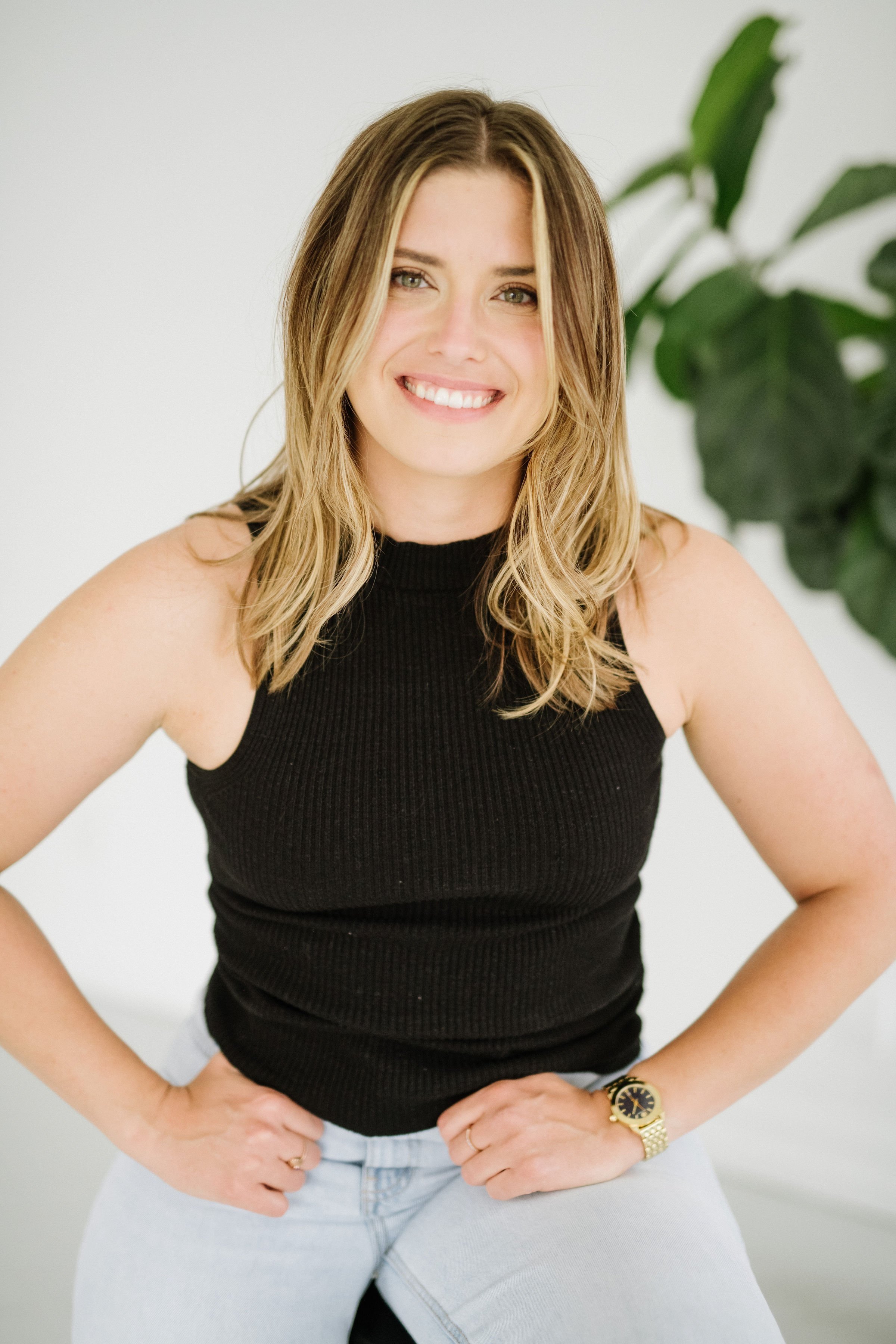 A young woman with shoulder-length blonde hair, smiling, wearing a black sleeveless top, light blue jeans, and a gold watch, standing in front of a white background with a green plant.