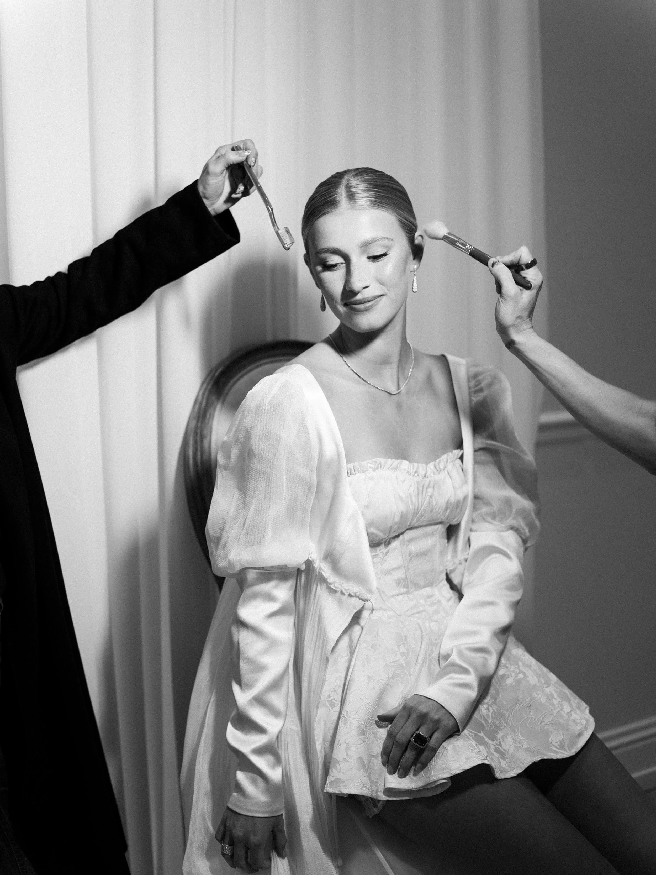 artistic shot of bride seated in a chair, with hands in either side reaching in to do her hair and makeup