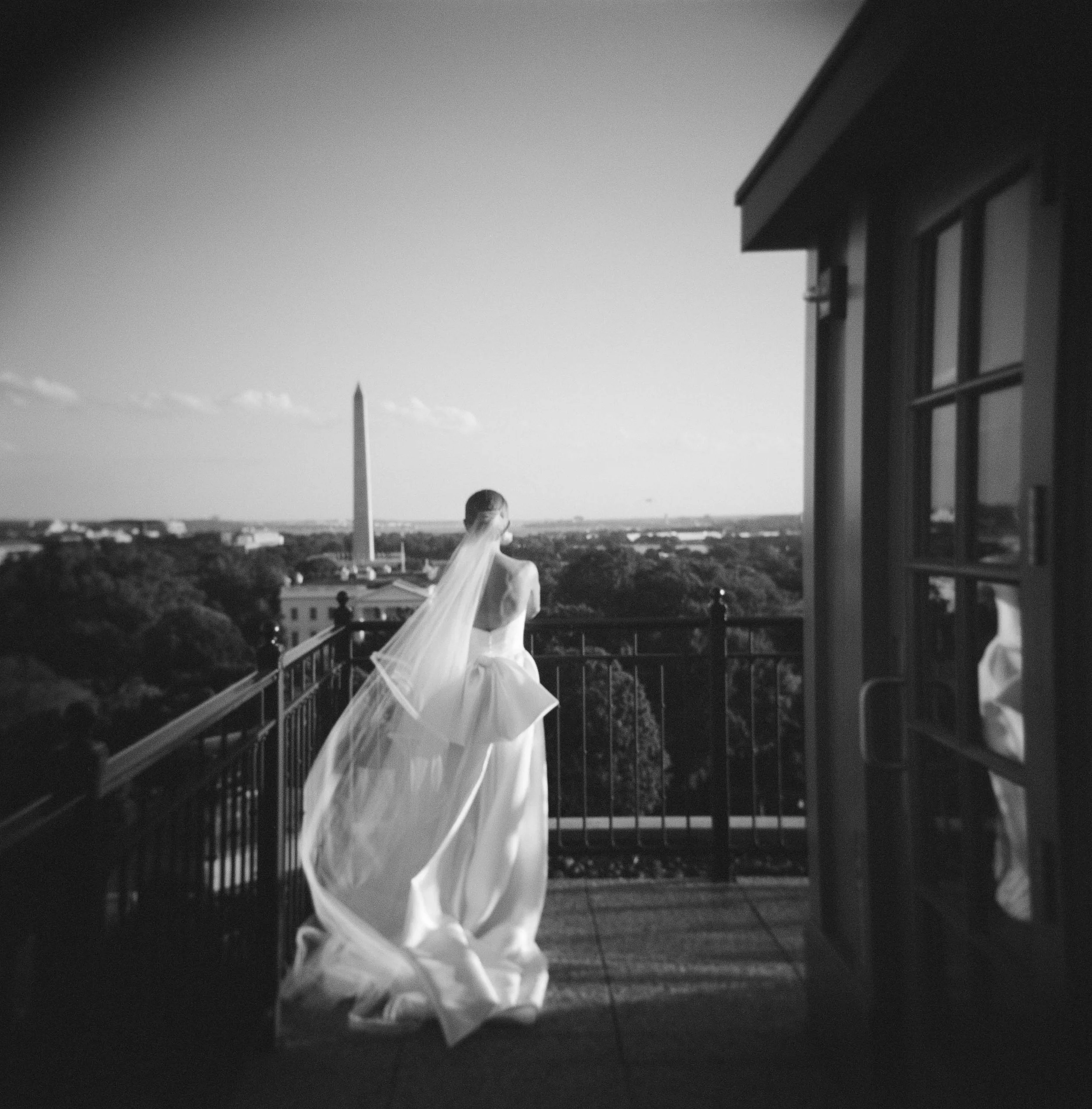 black & white film photo of a bride standing on a balcony in DC, facing the Washington monument