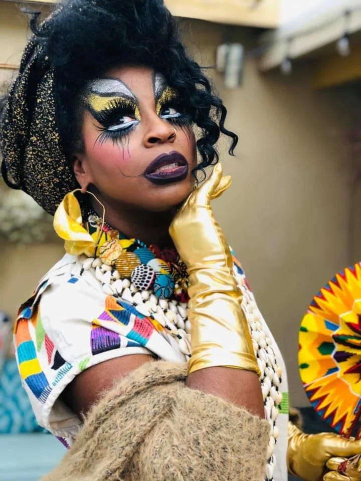 Beatrice Thomas, aka Black Benatar, looks over their right shoulder with a gold gloved hand under their chin.