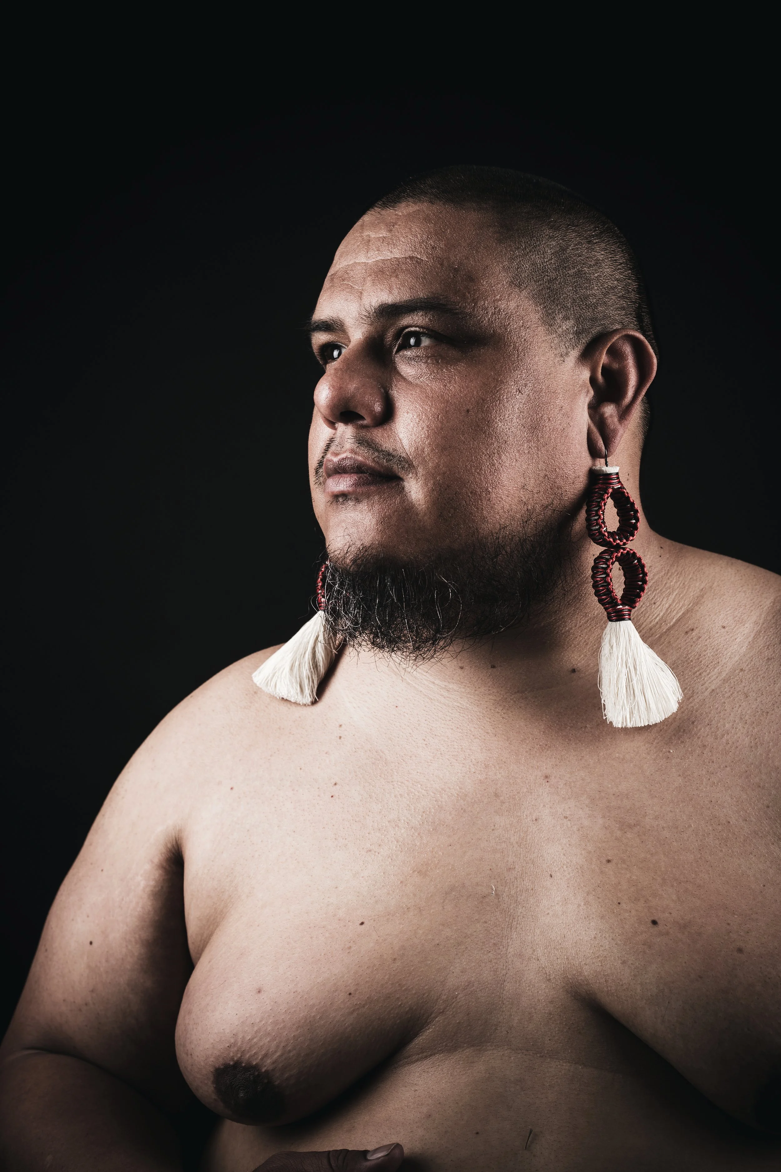 Dorian Wood, softly lit in front of a black background, looks off calmly to their right wearing no shirt and long red beaded earrings with white tassels.