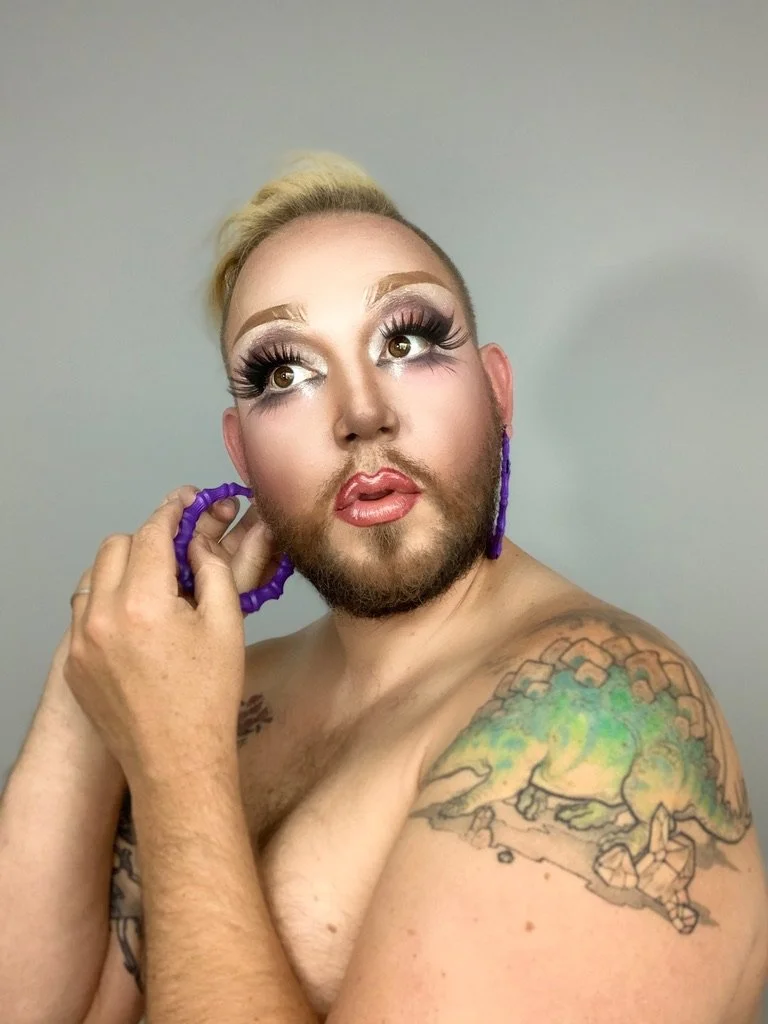 Headshot of Kevin Seaman in drag makeup, shirtless, putting on purple hoop earrings