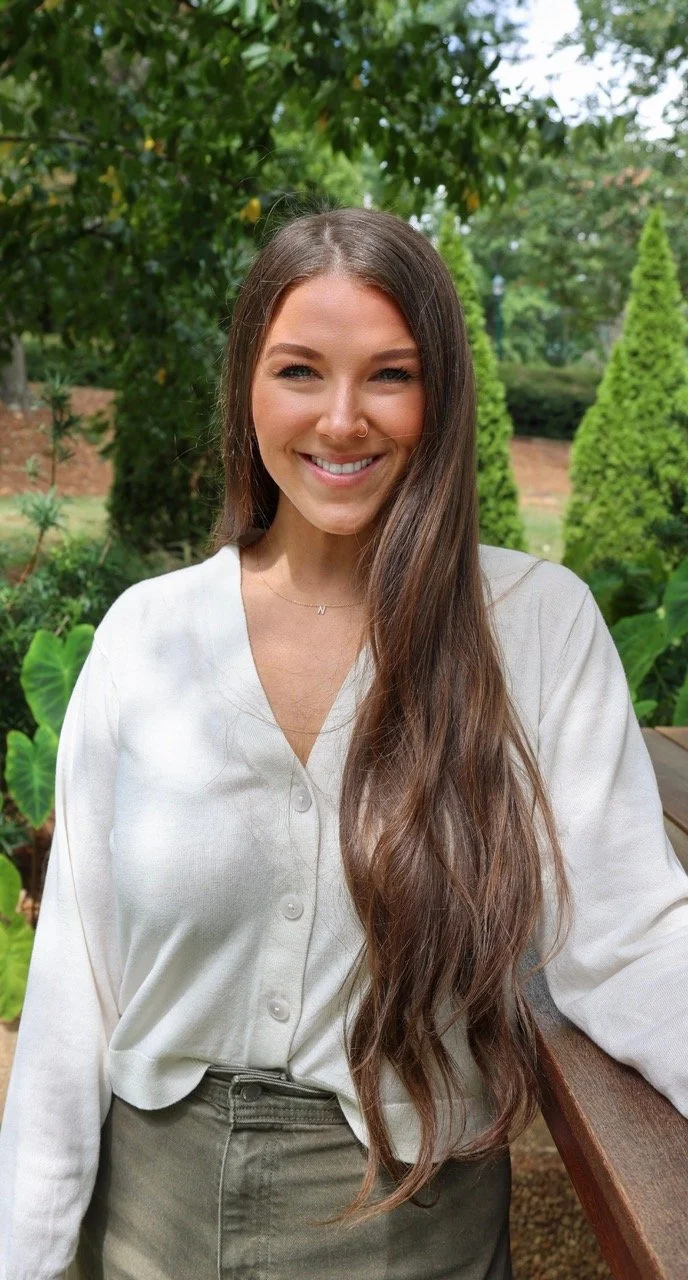 Nikki McCrimmon, a postpartum doula is a caucasian women with long brown hair smiling a warm and inviting smile and standing in front of trees outdoors with sunshine on her face.