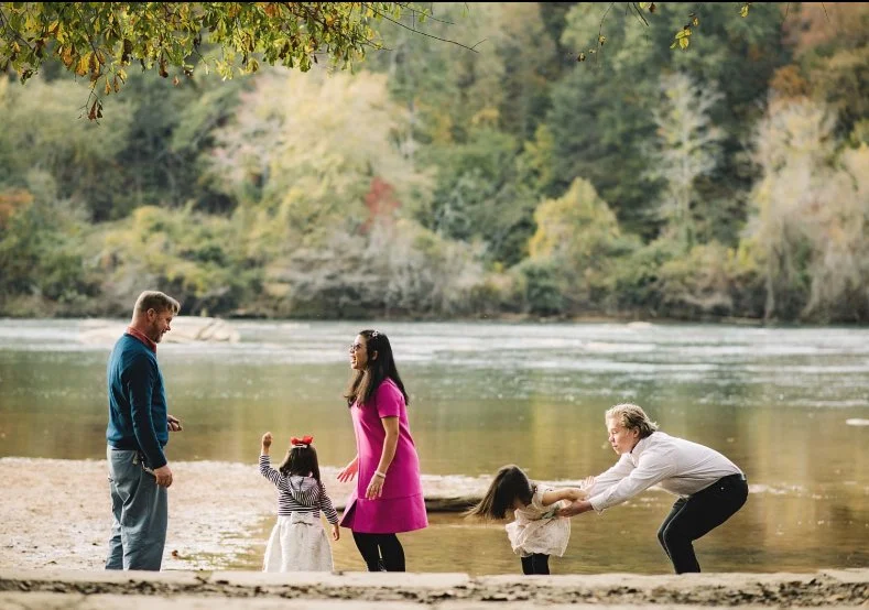 A family of five at the riverbank during autumn, with trees in fall colors in the background; two adults and three children playing and interacting.