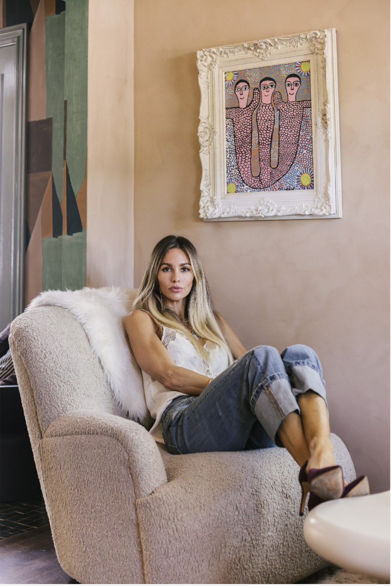 A woman sitting on a beige plush armchair in a room with pastel-colored walls, decorated with abstract artwork and framed colorful folk art with three figures and sun motifs.