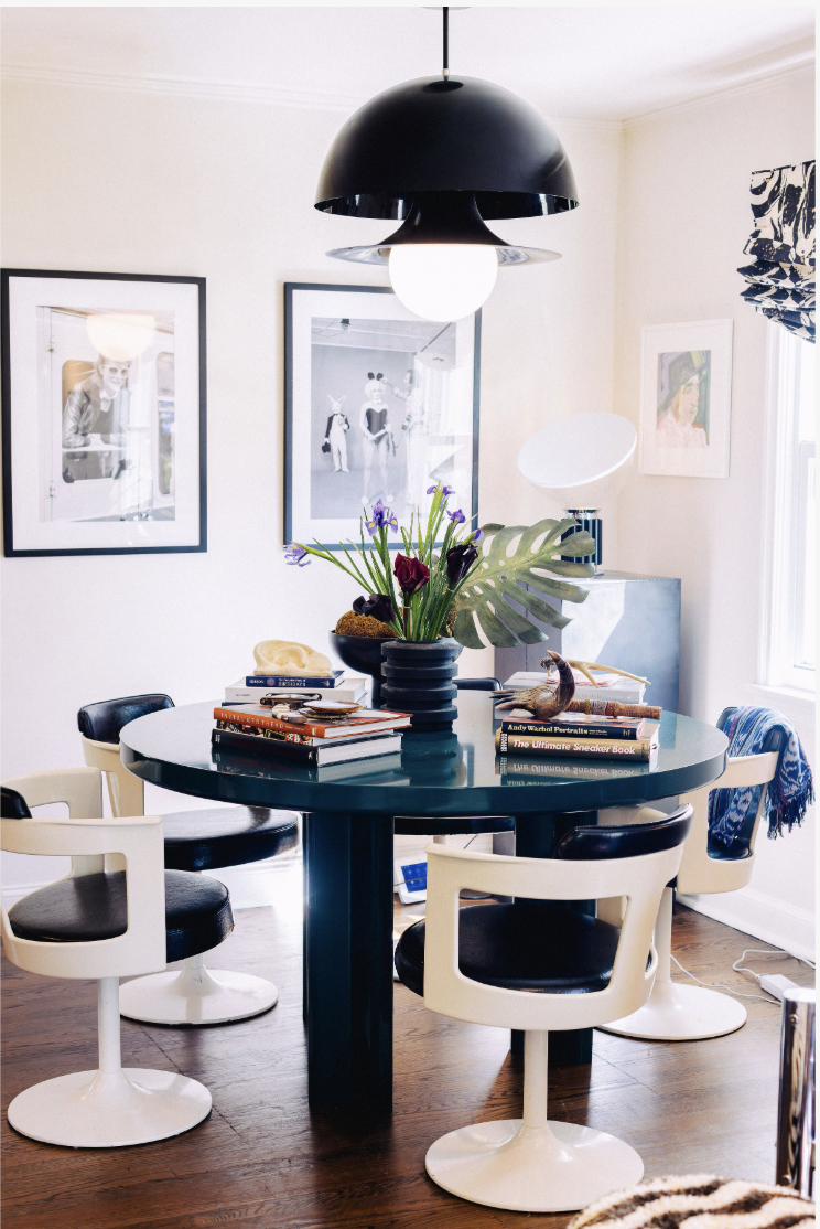 A modern dining room with a round dark blue table and seven chairs, with a large black pendant light hanging above, and framed artwork on the white walls.
