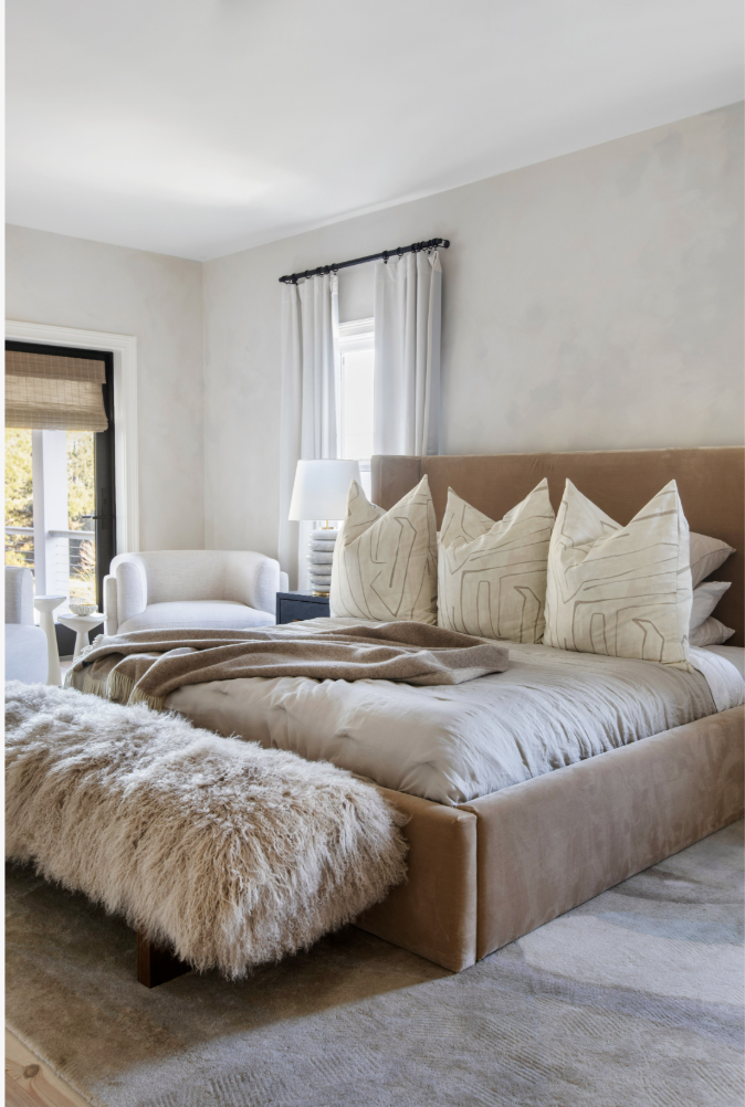 A cozy bedroom with a large bed featuring beige upholstered headboard and decorative pillows, a faux fur blanket, a nightstand with a lamp, and a sitting area with armchairs by a sliding door and window with white curtains.
