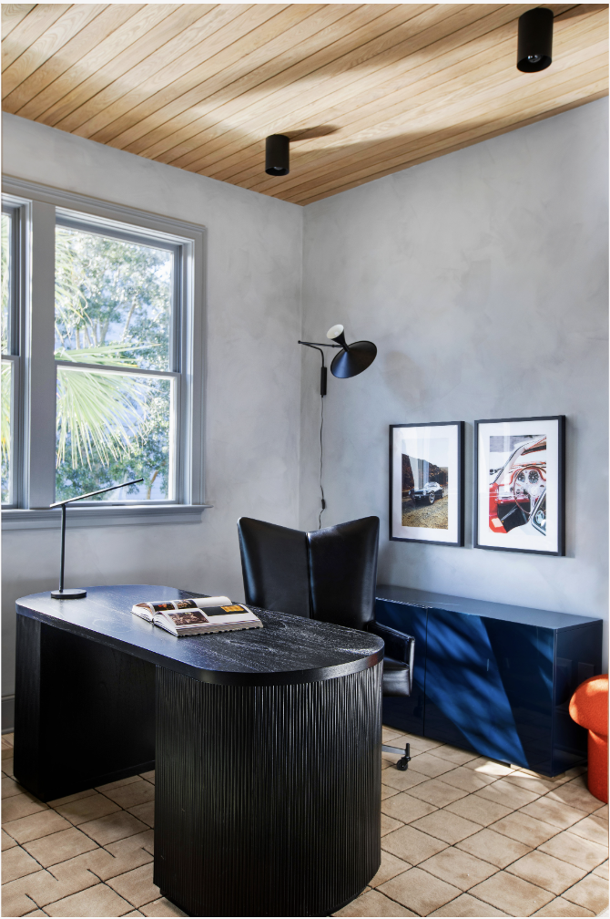 Home office with black desk, black leather chair, wall-mounted lamp, framed pictures, window with greenery outside, wooden ceiling, tiled floor, blue cabinet, orange chair, and natural sunlight.