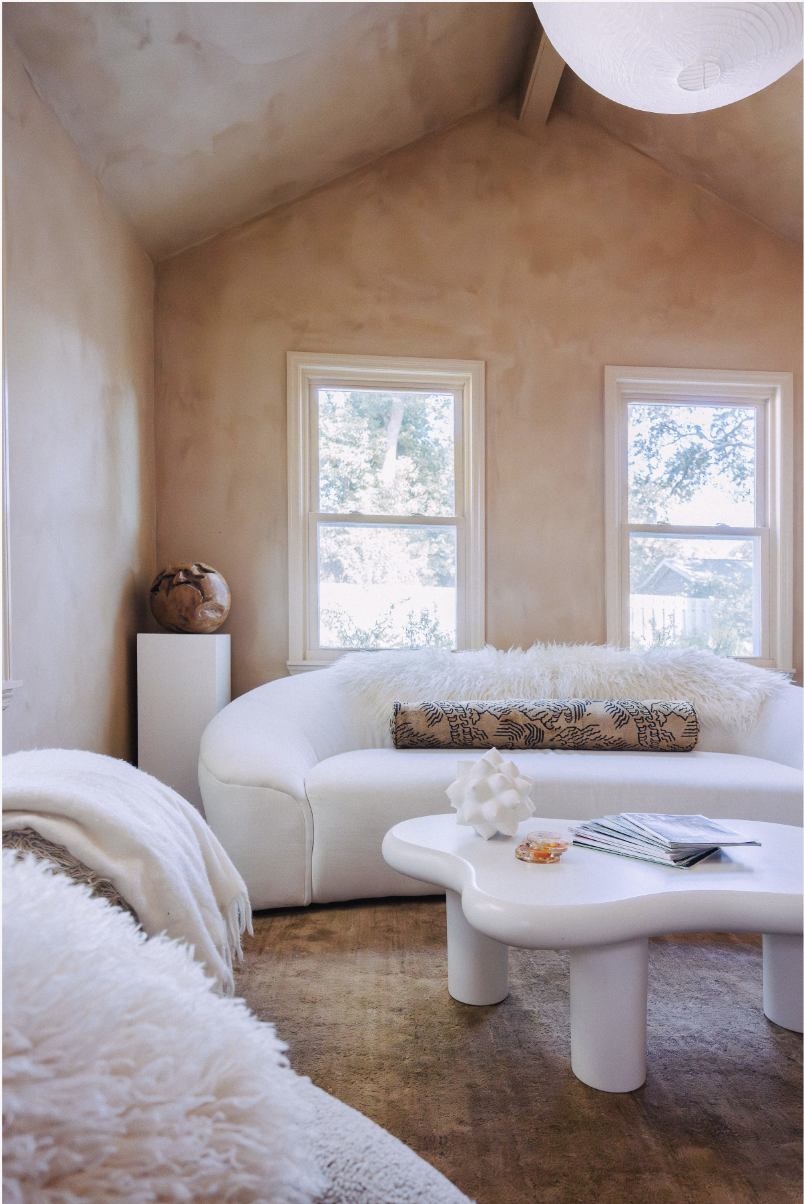 Living room with beige limewash textured walls, two windows letting in natural light, a white curved sofa with fluffy pillows, a white abstract coffee table with magazines and decorative items, and a corner vase on a small pedestal.