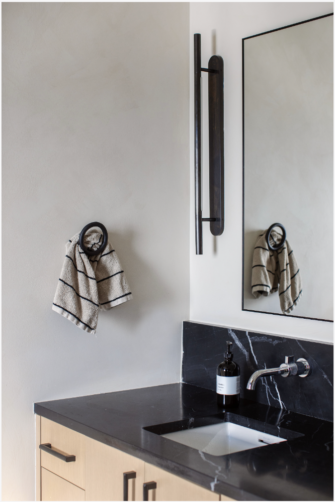 A modern bathroom wall with a beige towel hanging on a black ring, a black vertical wall hook, a mirror, a black soap dispenser, a black marble sink countertop, and a cabinet with black handles.