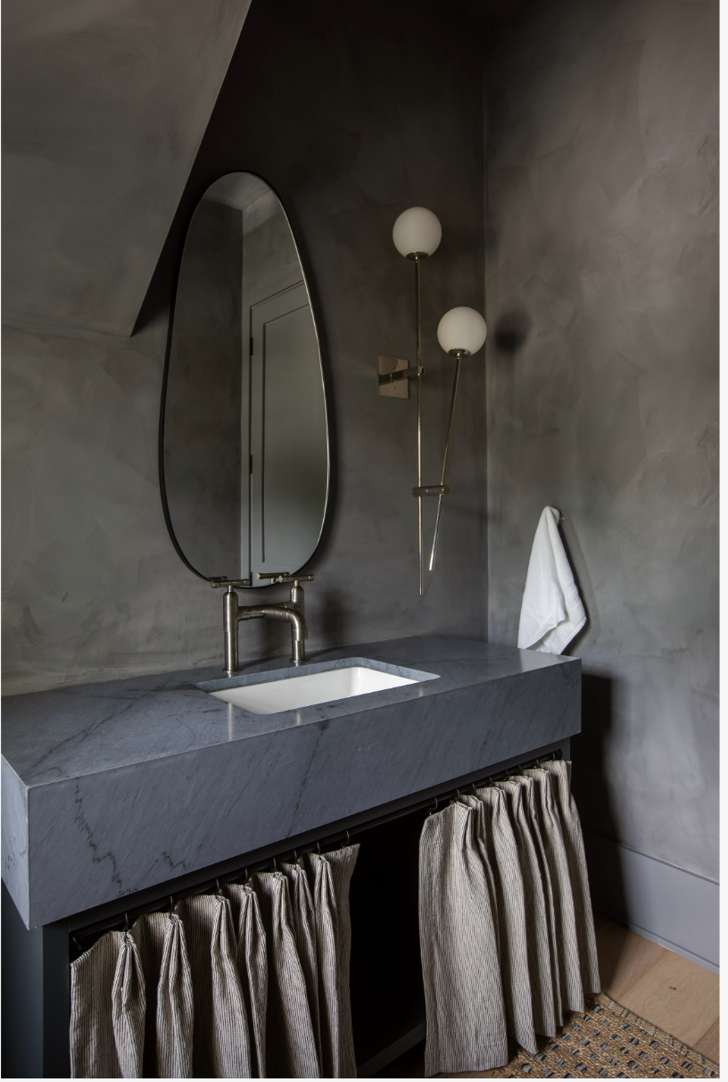 A modern bathroom vanity with a marble countertop, an oval mirror, two globe wall-mounted light fixtures, a white towel hanging on the wall, and beige curtains underneath the sink. The walls are dark gray with a textured limewash finish.