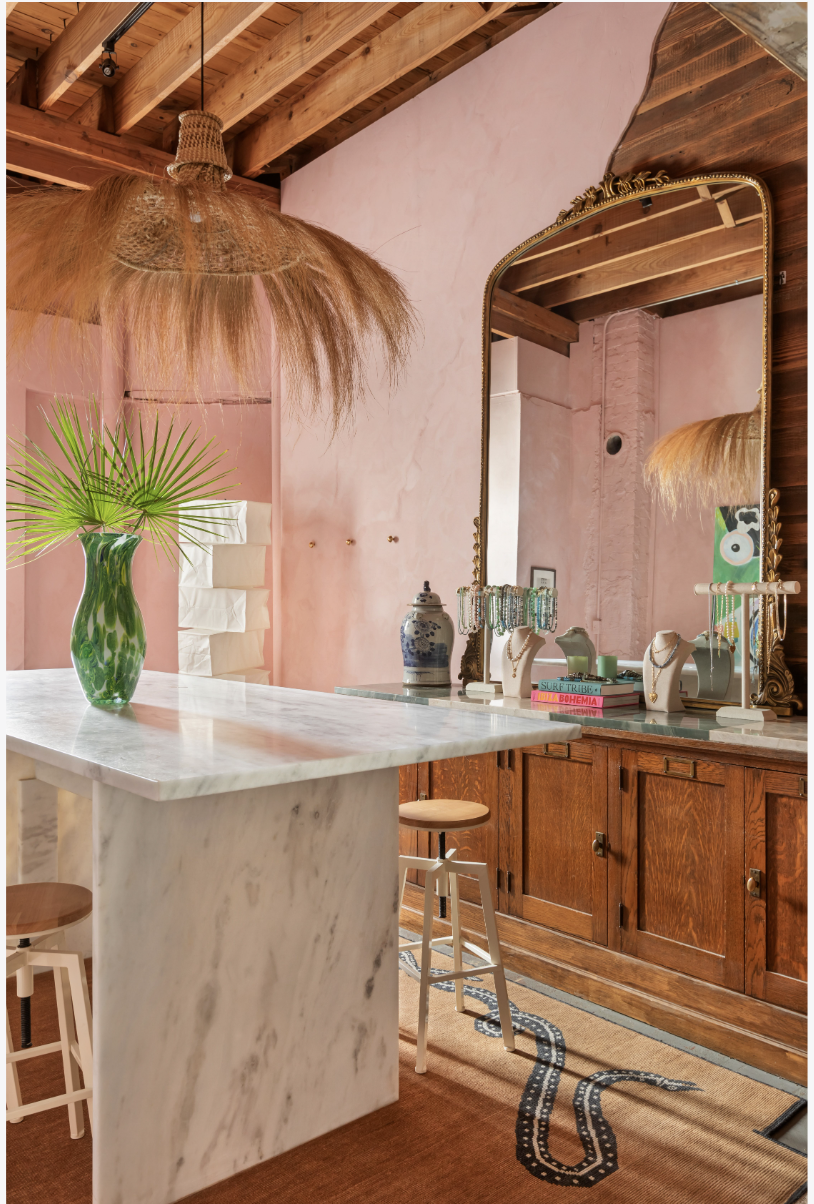A room with a pink wall and wooden ceiling beams, featuring a white marble countertop with a green vase holding palm leaves, a brass-framed mirror, various jewelry displays, a ceramic jar, and a stack of colorful books. There is a wicker pendant ligh