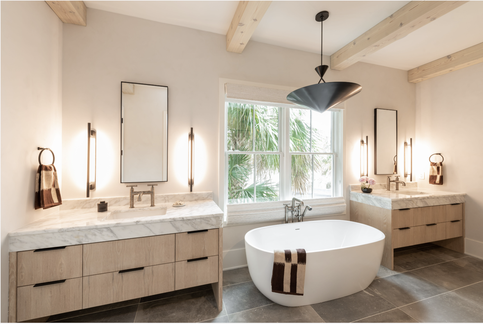 Modern bathroom with double vanities, marble countertops, rectangular mirrors, wall-mounted lights, a freestanding bathtub in front of a large window, and a wooden ceiling with exposed beams.
