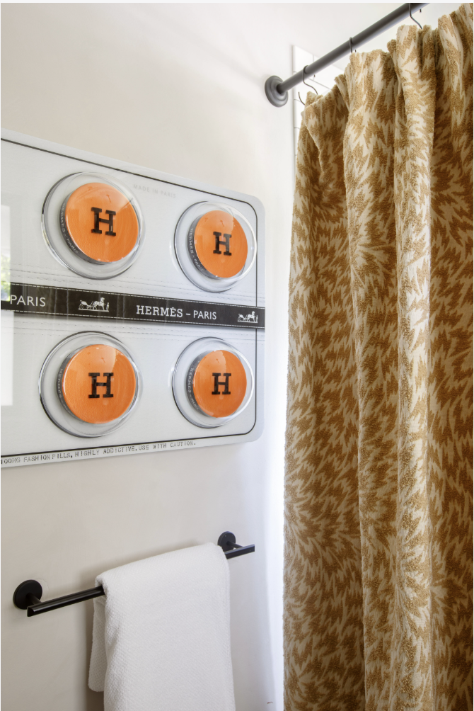 Wall-mounted towel rack with a white towel, and a decorative wall art piece with four orange circles marked with an 'H' in black, adjacent to a patterned gold and white curtain.