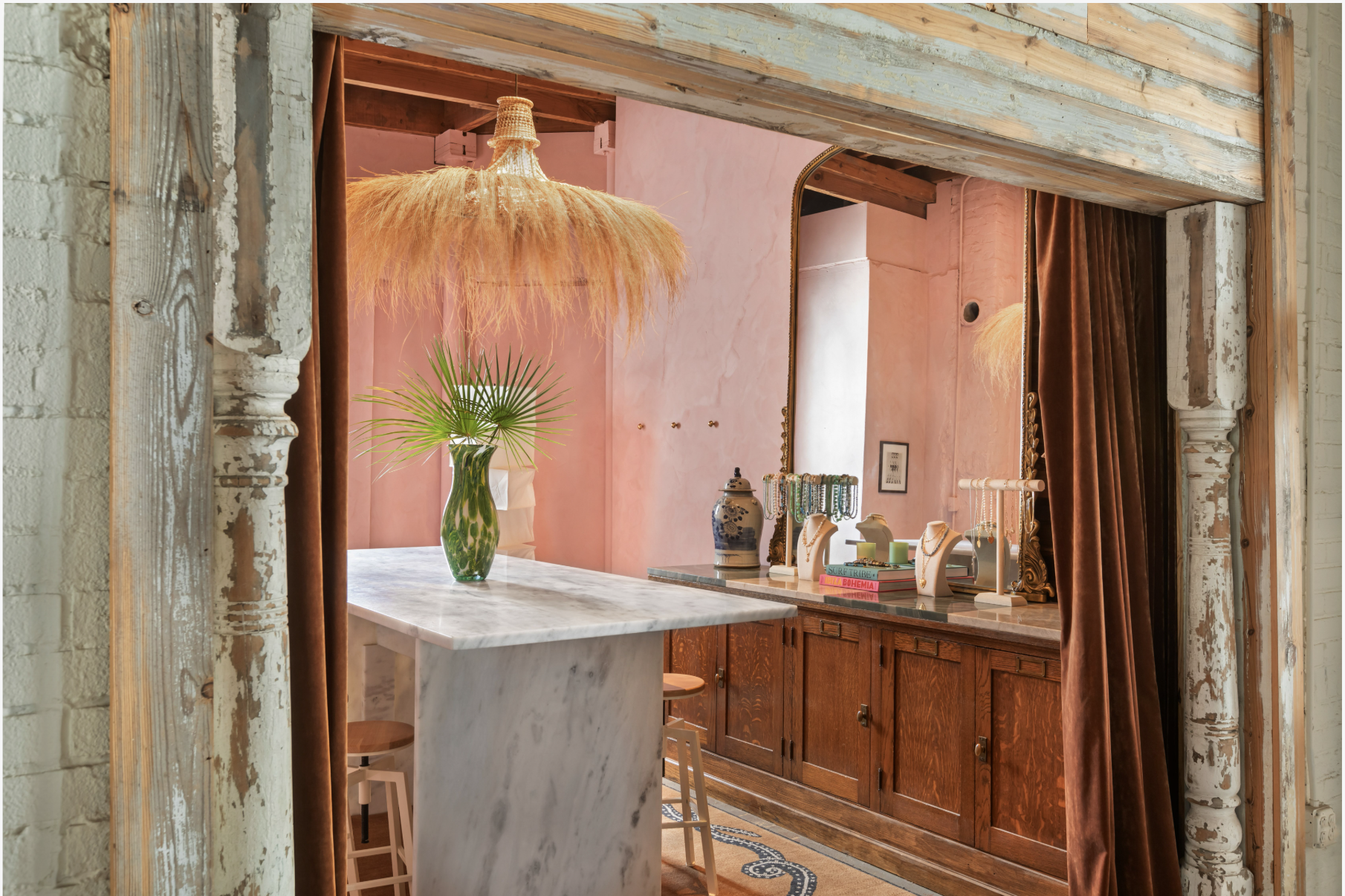 Decorative boutique interior seen through a rustic wooden and brick doorway with velvet curtains, a large mirror, a marble island with a vase and palm leaves, and jewelry displays on a wooden counter.