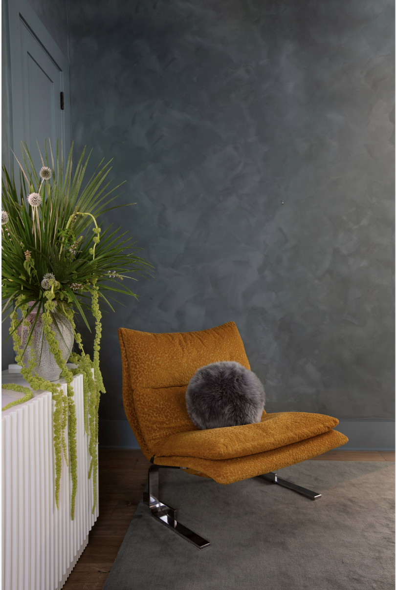 An orange lounge chair with a gray furry cushion in a room with a textured gray wall, a large floral arrangement in a white vase on a white ribbed side table, and a gray rug on wooden floor.