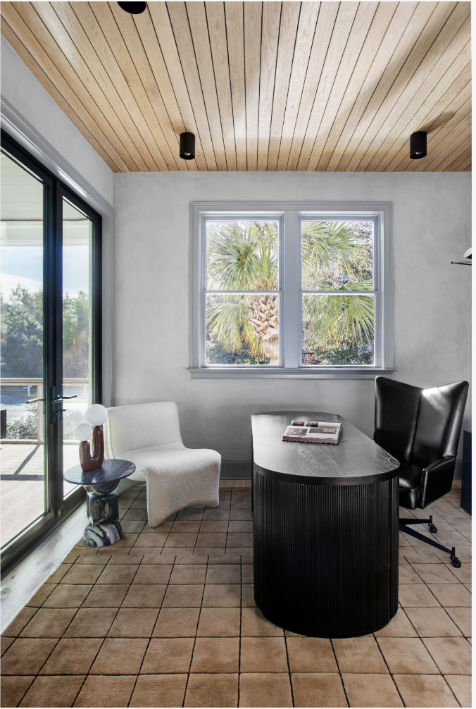 A modern office space with a black desk, black swivel chair, white armchair, and a side table with a decorative lamp. Large window with palm trees outside, and a sliding glass door leading to a balcony. Wooden ceiling and tiled floor.