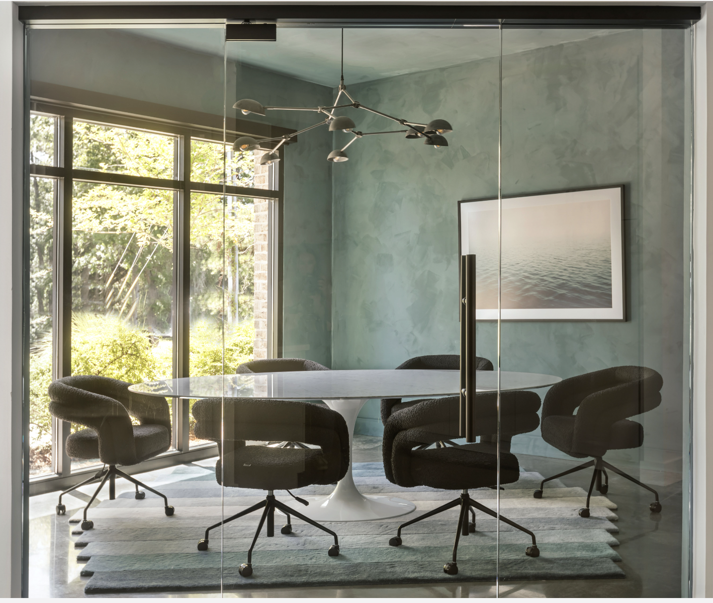 Modern conference room with glass walls, round table, six black swivel chairs, large windows, green walls, artwork of water, and a ceiling light fixture.