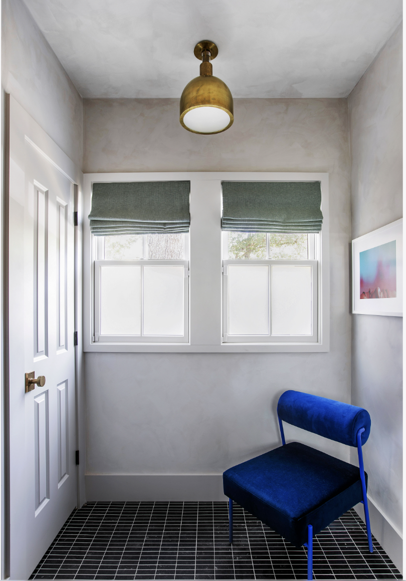 A small room with two windows, green blinds, and a gold ceiling light, containing a blue velvet chair with thin blue legs and a framed picture on the wall.