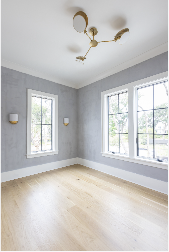 Empty room with two large windows, light wood flooring, gray walls with white trim, and a gold modern ceiling light fixture.
