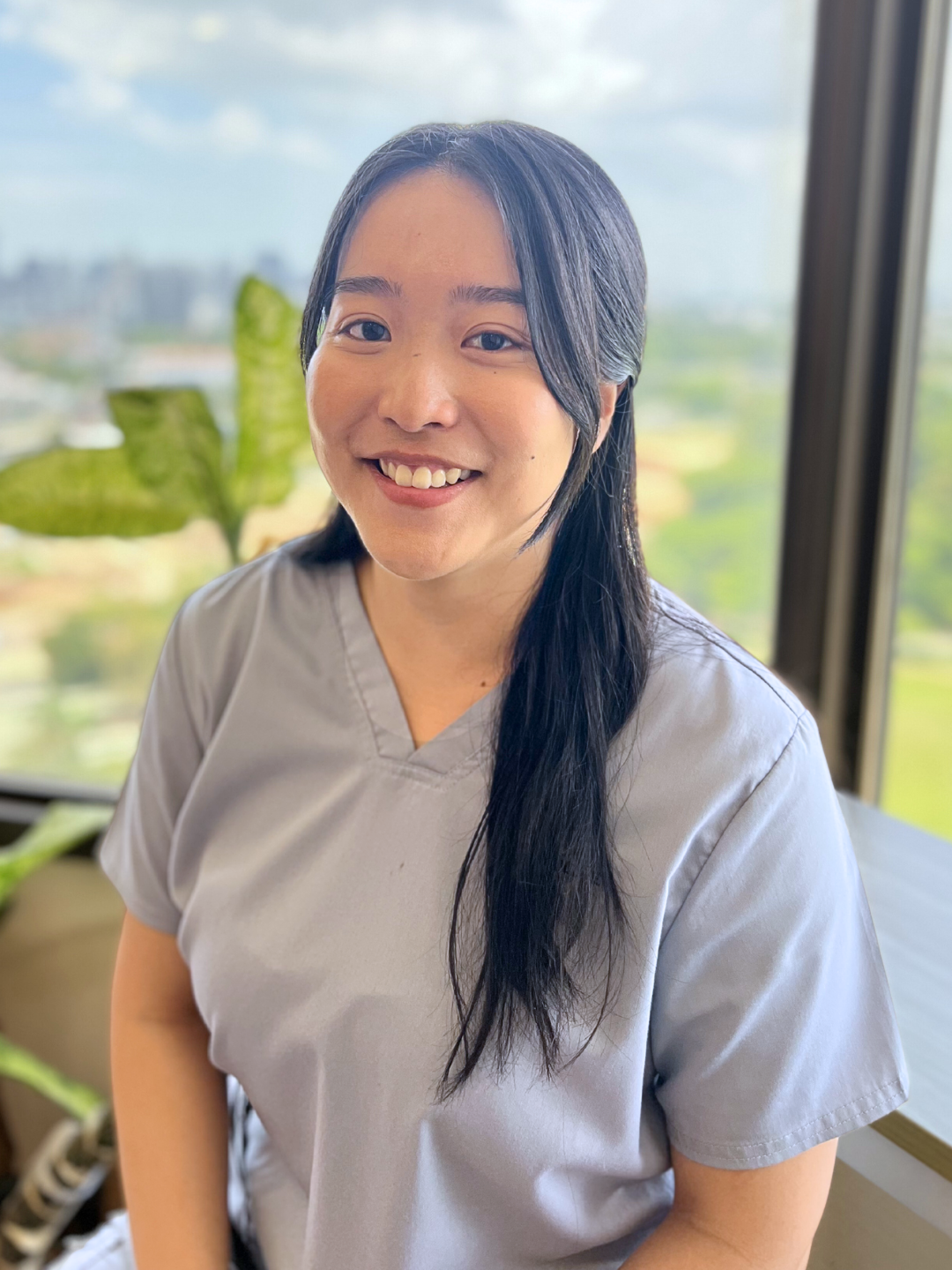 A woman with long black hair and a gray medical scrub top smiling inside an office with large windows and a view of trees and buildings in the background.