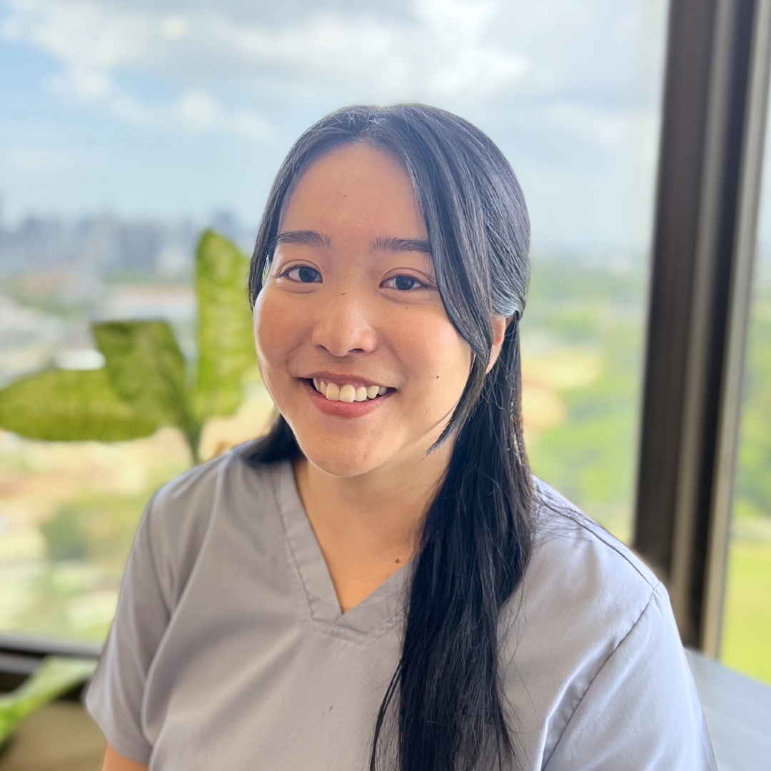 Smiling woman with long black hair wearing gray scrubs, standing in front of large window with green plants and city landscape outside.