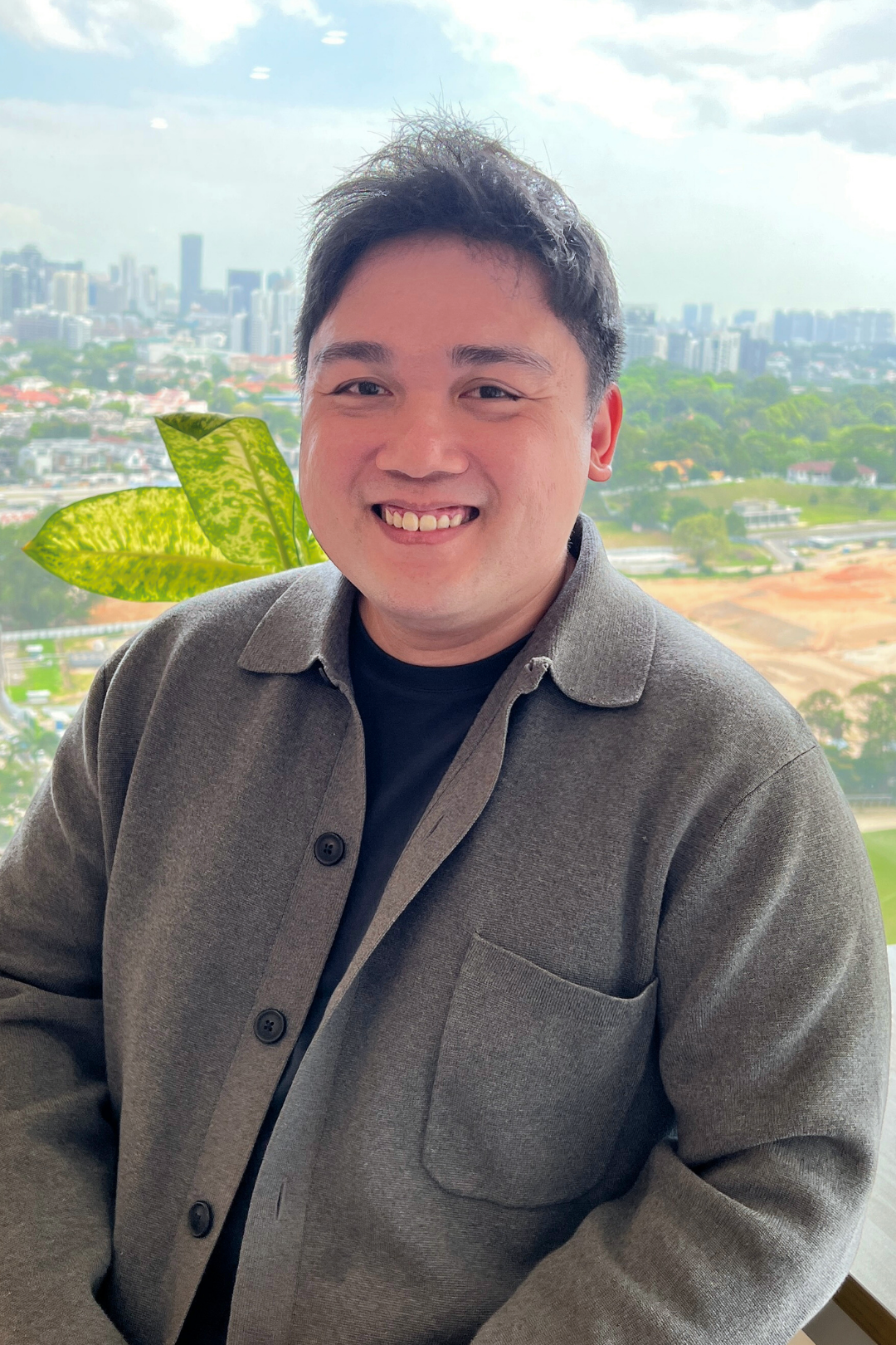 A smiling man with dark hair, wearing a gray jacket and black shirt, standing in front of a large window with a cityscape view of tall buildings, greenery, and an overcast sky.