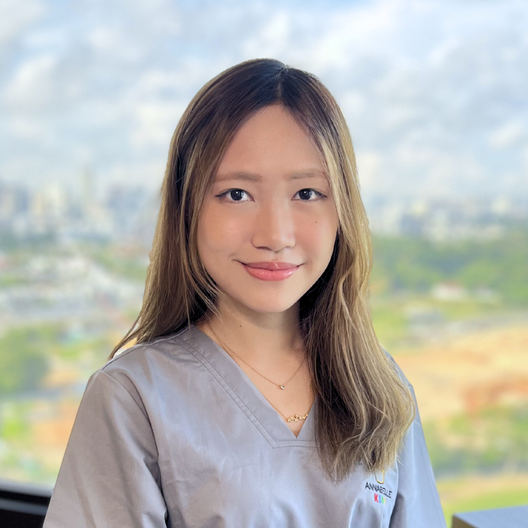 A young woman with long, wavy brown hair, smiling, wearing gray scrubs with a name tag that says Annabelle, standing in front of a window with a cityscape and cloudy sky in the background.