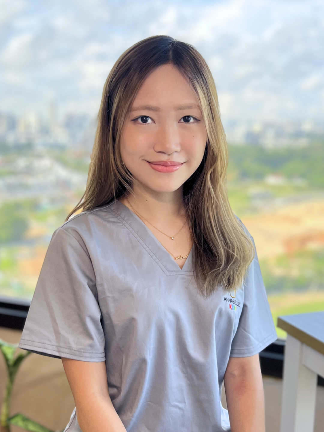 A young woman with light brown hair and a light gray uniform shirt with a name tag that says "Annabelle" standing indoors near a window with a city landscape outside.