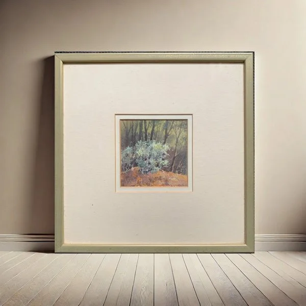 Framed miniature by Margaret Wills shown in softly lit interior with neutral wall and timber shelf. Artwork features sparse wattle bushes in pastel hues on cream paper.