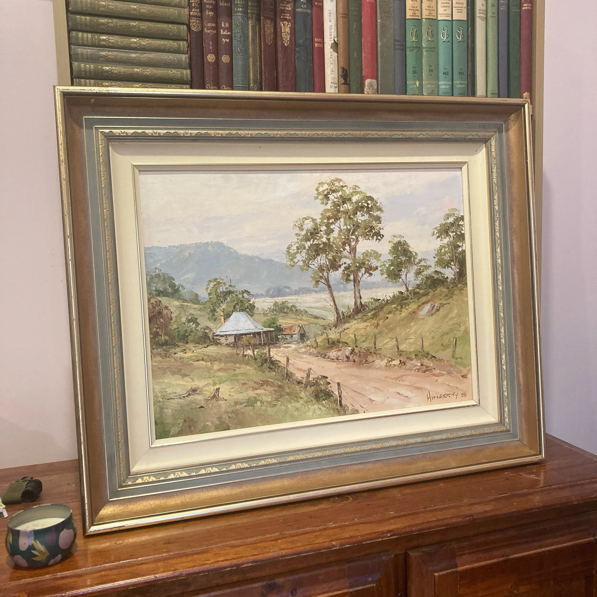 Framed oil painting by John Hingerty titled "Hut from Araluen," showing a country farmhouse and barn on a dirt road, surrounded by trees and hills in pastel tones.