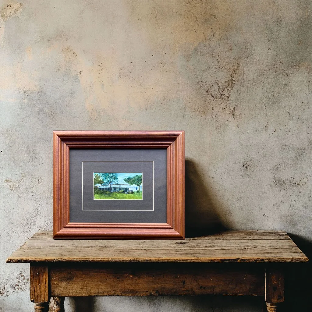 Framed miniature painting by John Thiessen depicting a white-roofed house among greenery, shown with double mat and timber frame in interior setting
