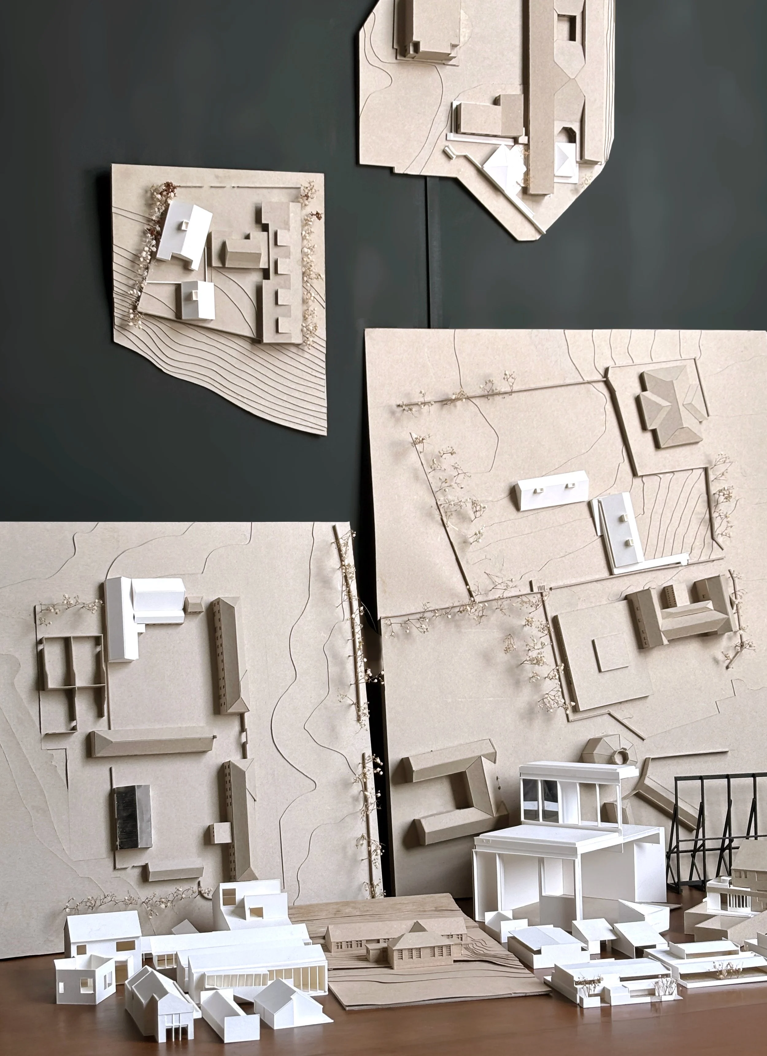 Architectural models of buildings and urban layouts displayed on a table with large topographical maps in the background.