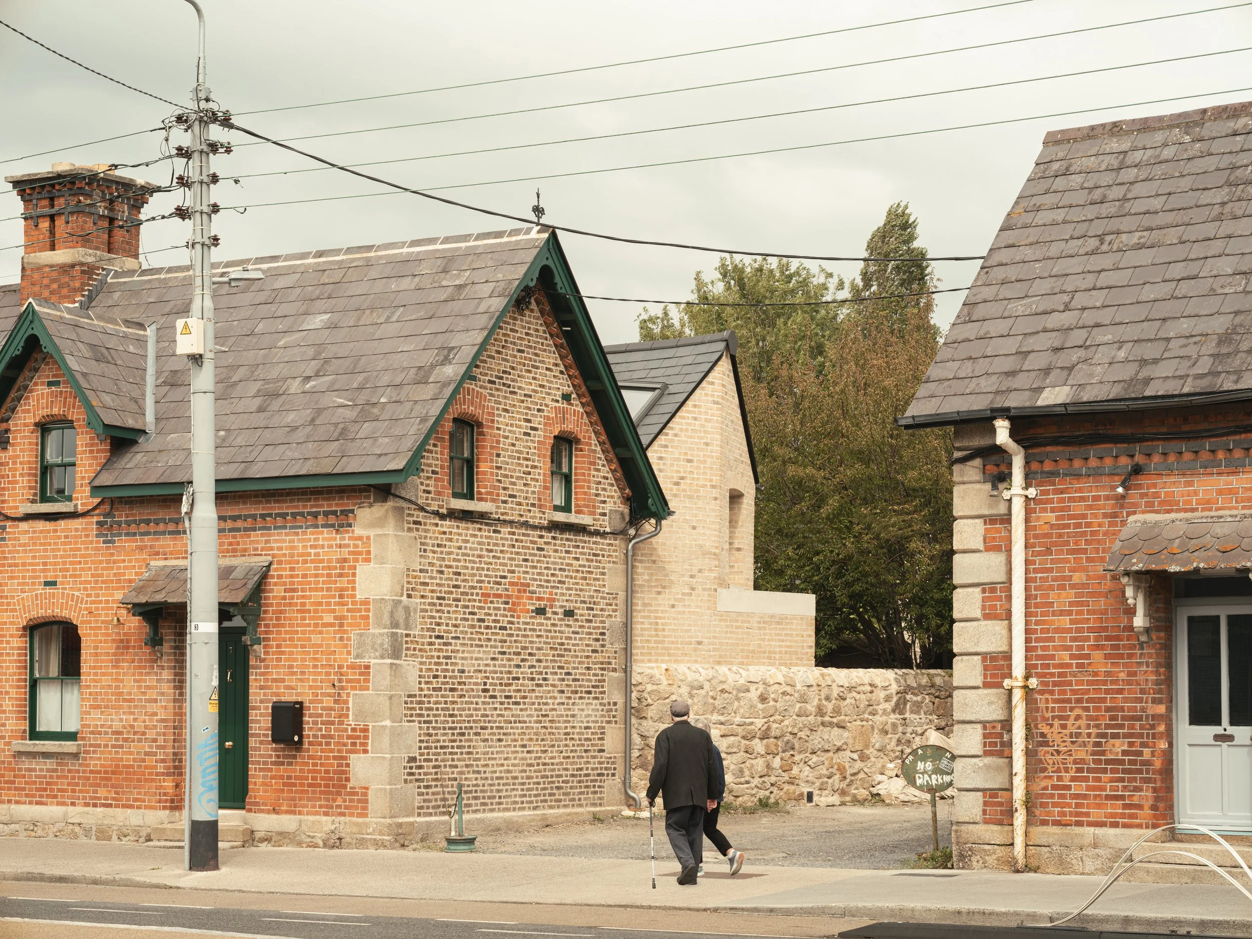 A man in a black coat and cap walking with a cane in front of brick buildings on a street. There are overhead power lines and a pole, with a gray sky overhead.