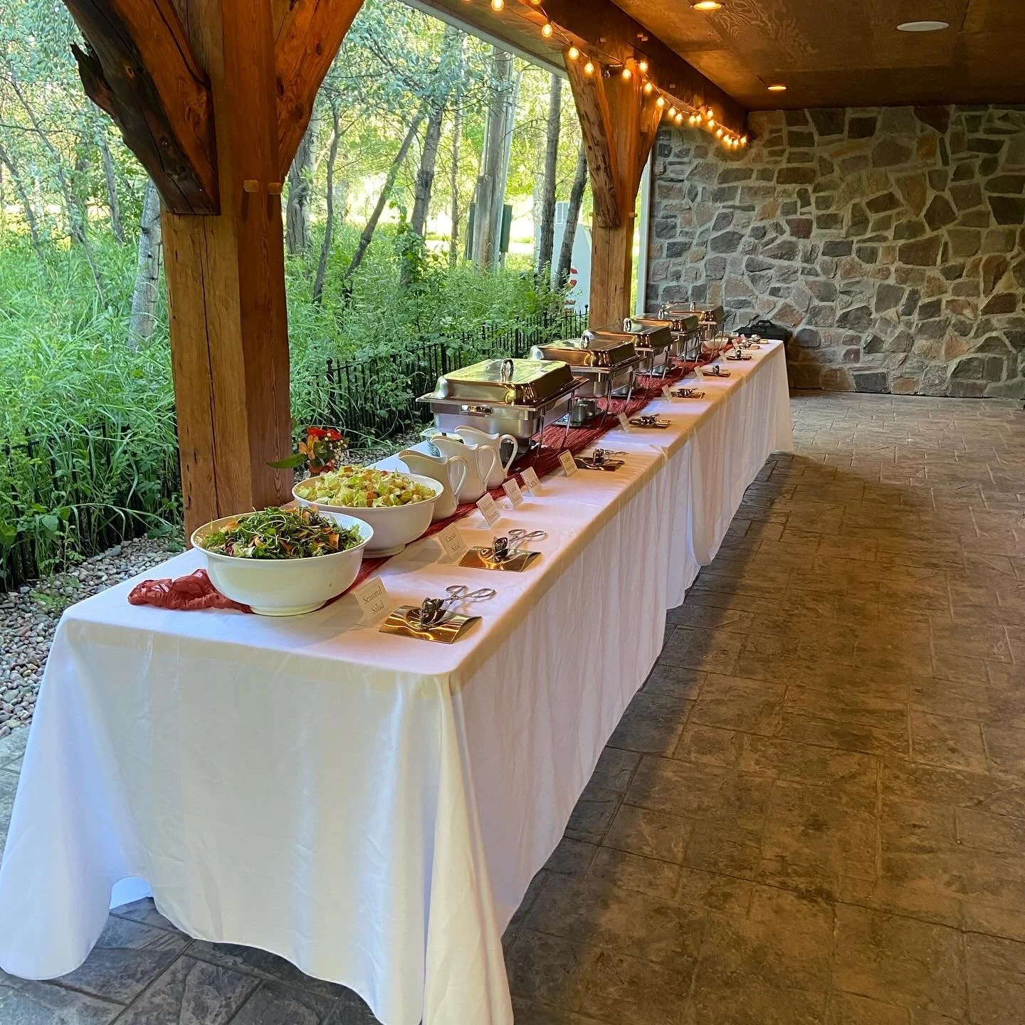 A long banquet table set for a buffet with chafing dishes, bowls of salad, and plates with clothing labels, in a rustic venue with wood beams, stone wall, string lights, and large windows showing greenery outside.