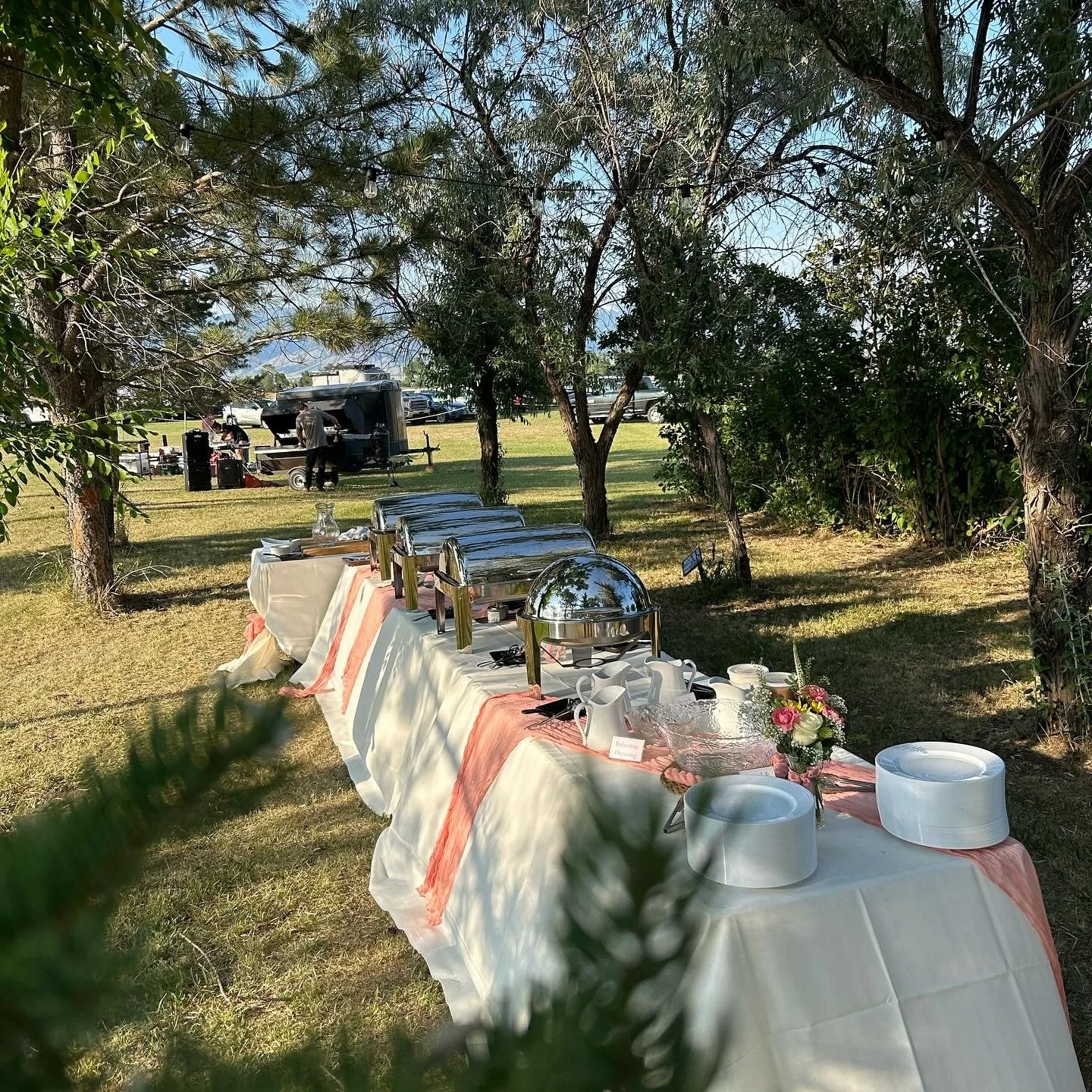 Outdoor buffet table with chafing dishes and tea set in a shaded garden area with trees, string lights, and parked cars in the background.