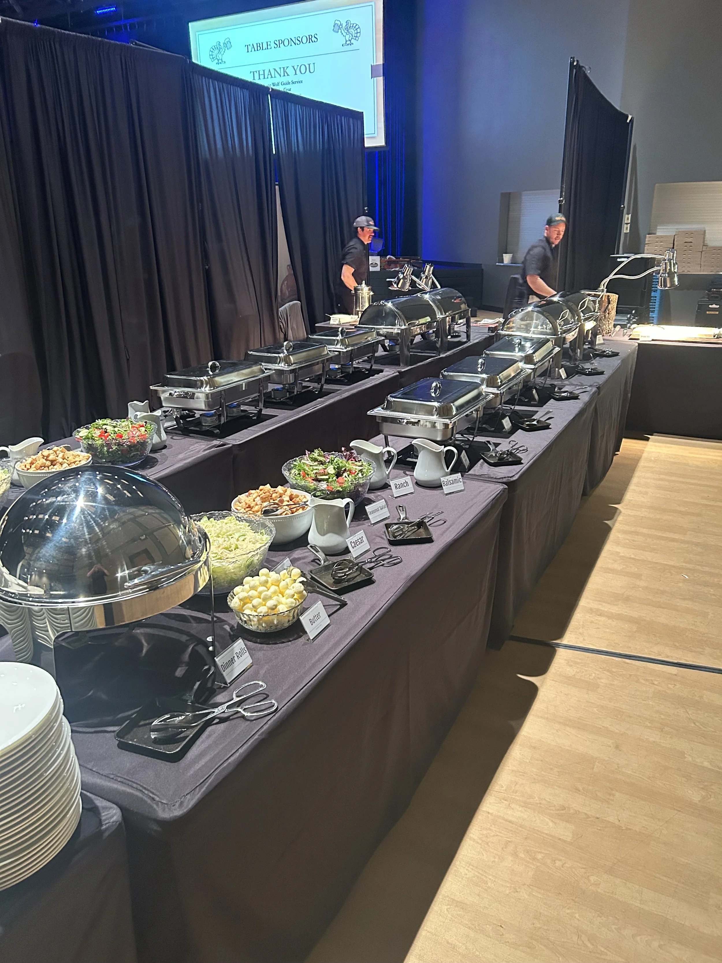 Buffet table with various dishes including salad, dinner rolls, and cheese, with chafing dishes and utensils, set in a banquet hall with black curtains and a sign overhead.