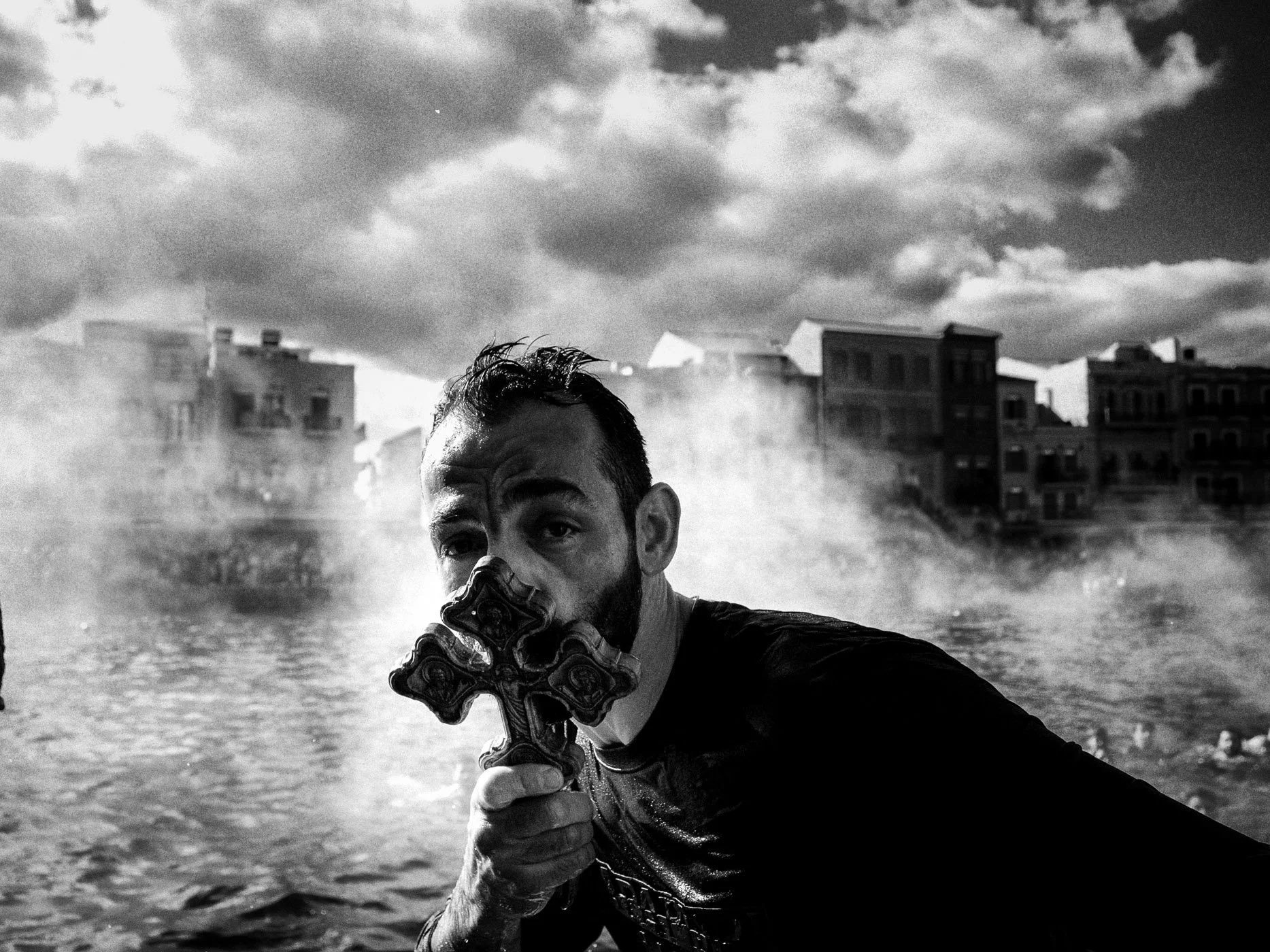 Man holding decorative cross near water, with mist and buildings in the background, black and white photo.