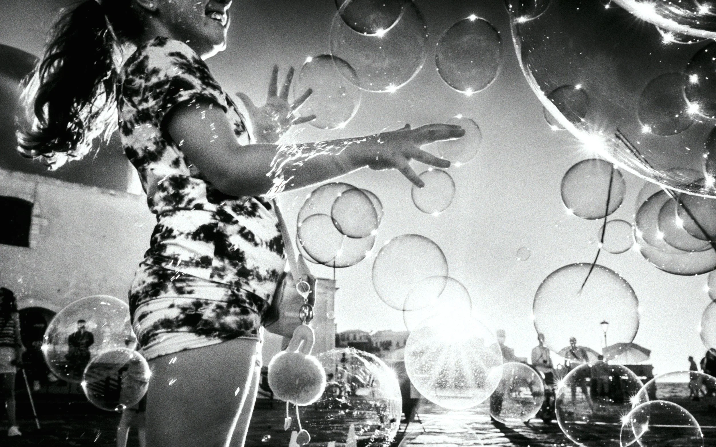 A young girl surrounded by large soap bubbles in a sunlit outdoor setting, wearing a tie-dye shirt. The image is in black and white, capturing her joyful expression.
