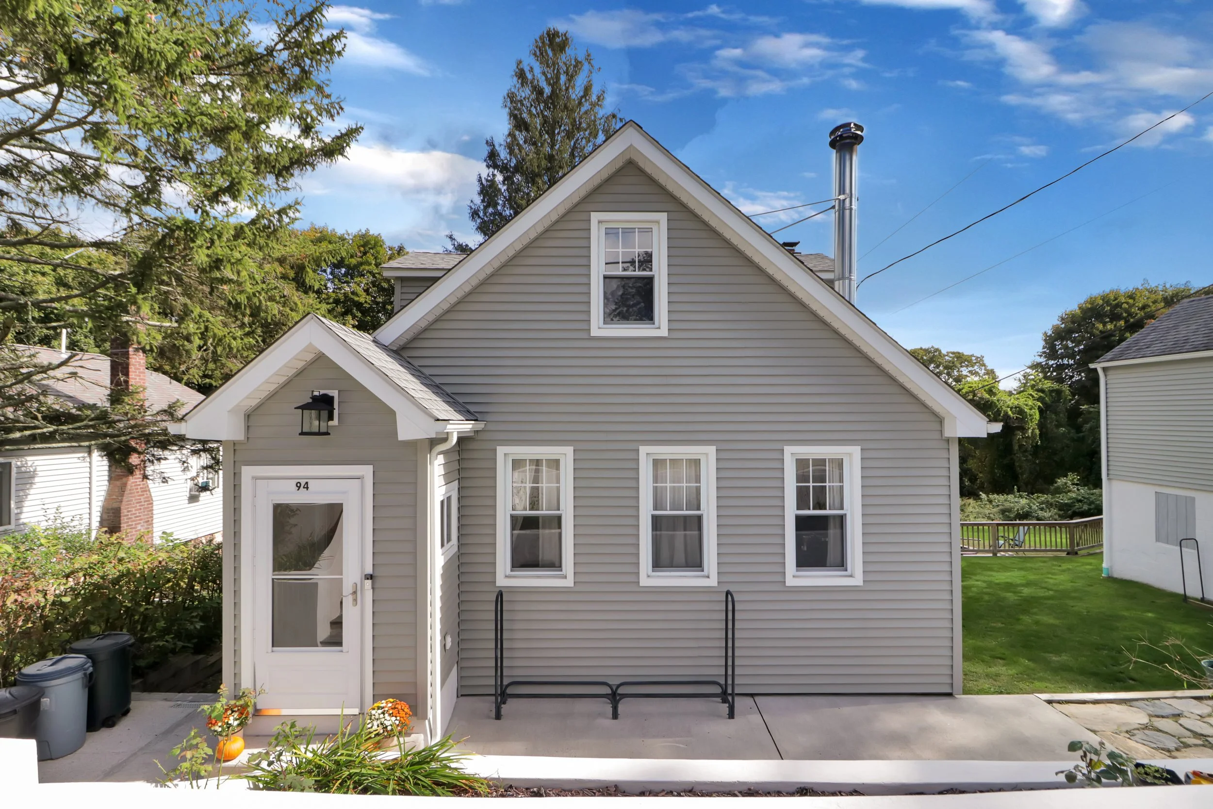 Front view of a gray house with white trim, featuring a small covered porch with a door labeled "94," three front windows, a chimney, and small landscaped area with flowers and pumpkins.