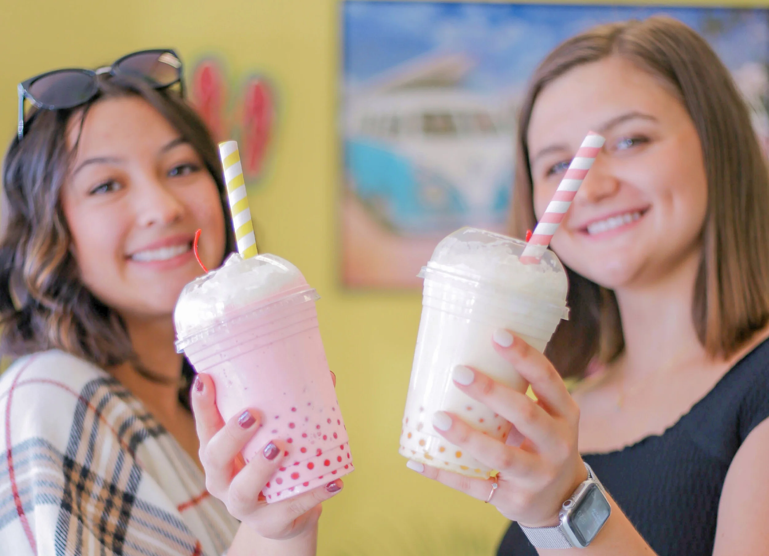 Two women each holding a bubble shake. One is light pink and one is light yellow. You can see some of the boba balls in the bottom of each shake.