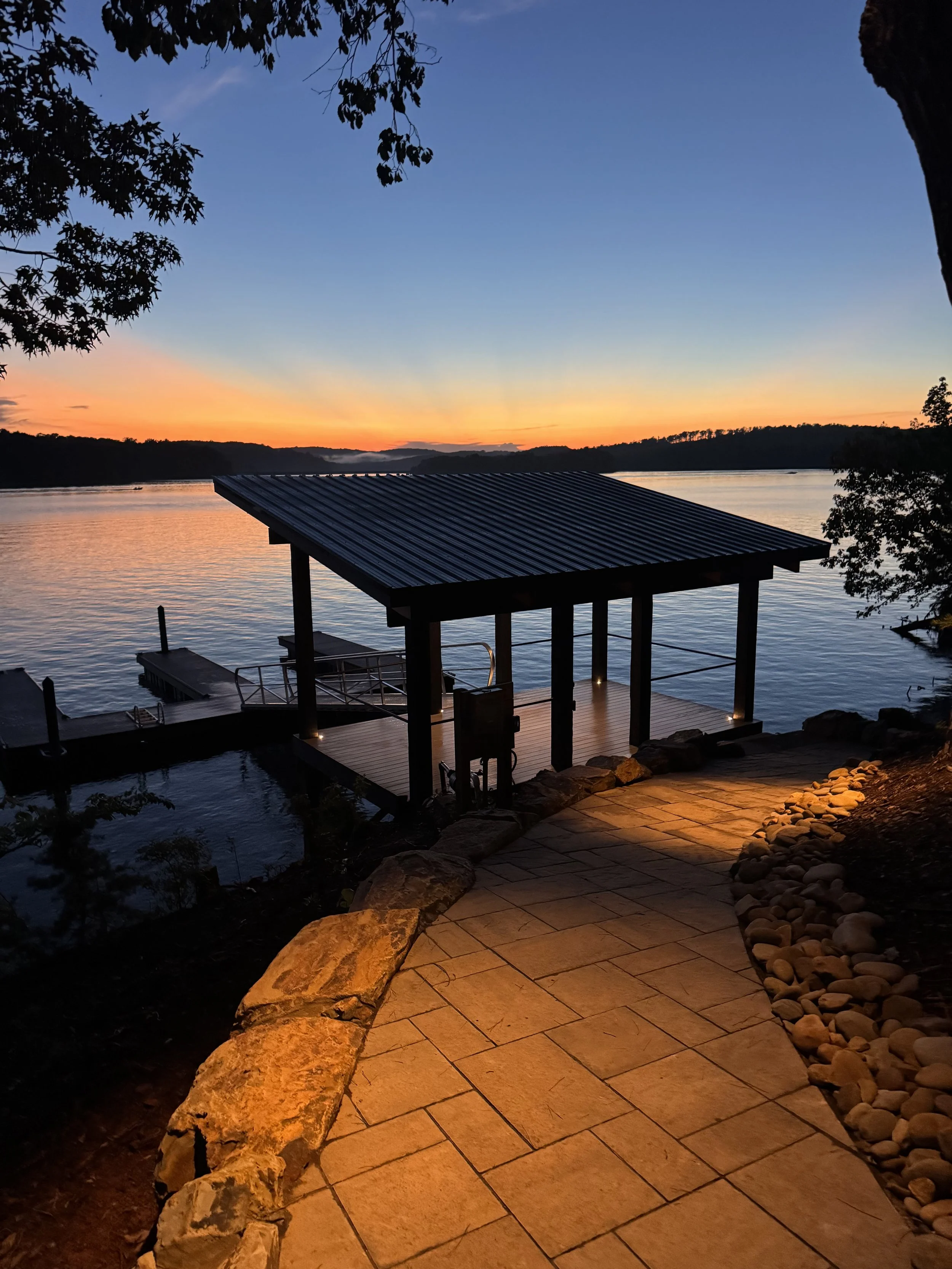 A lakeside scene at sunset with a wooden dock and covered structure, surrounded by trees and rocks, and warm outdoor lighting.