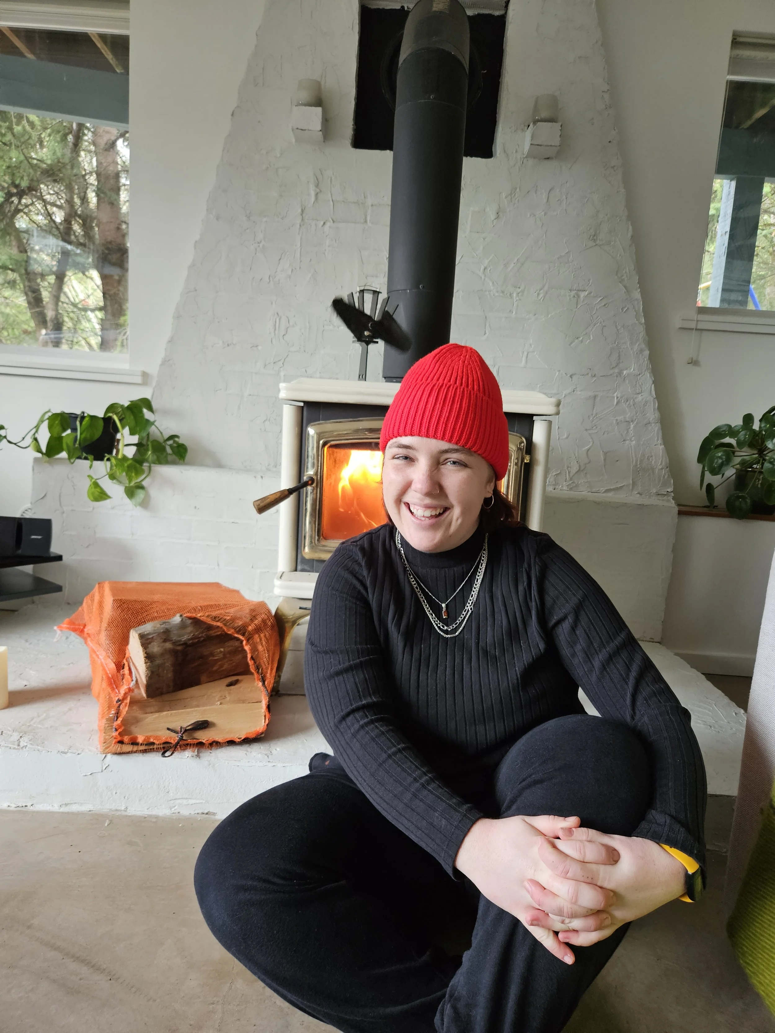 Trauma counsellor for 2SLGBTQIA+ online in BCg young woman wearing a red beanie and black sweater, sitting on the floor near a lit fireplace in a cozy room with houseplants and windows.