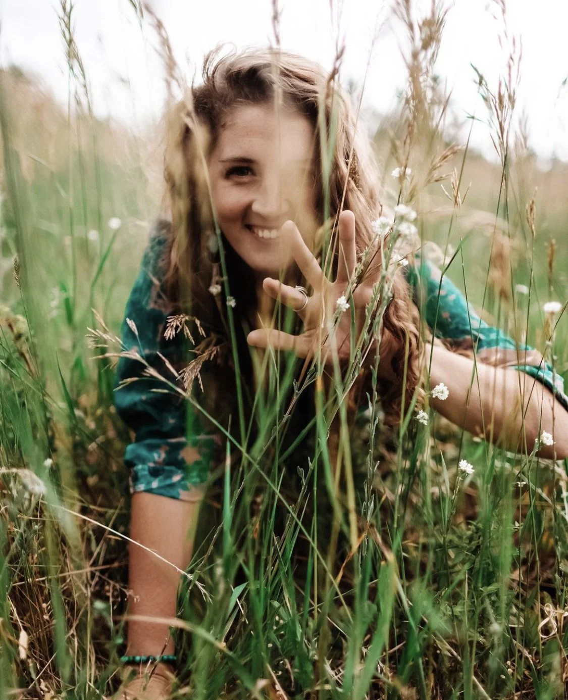 Woman with long hair smiling and making a peace sign while lying down in a grassy field with wildflowers.