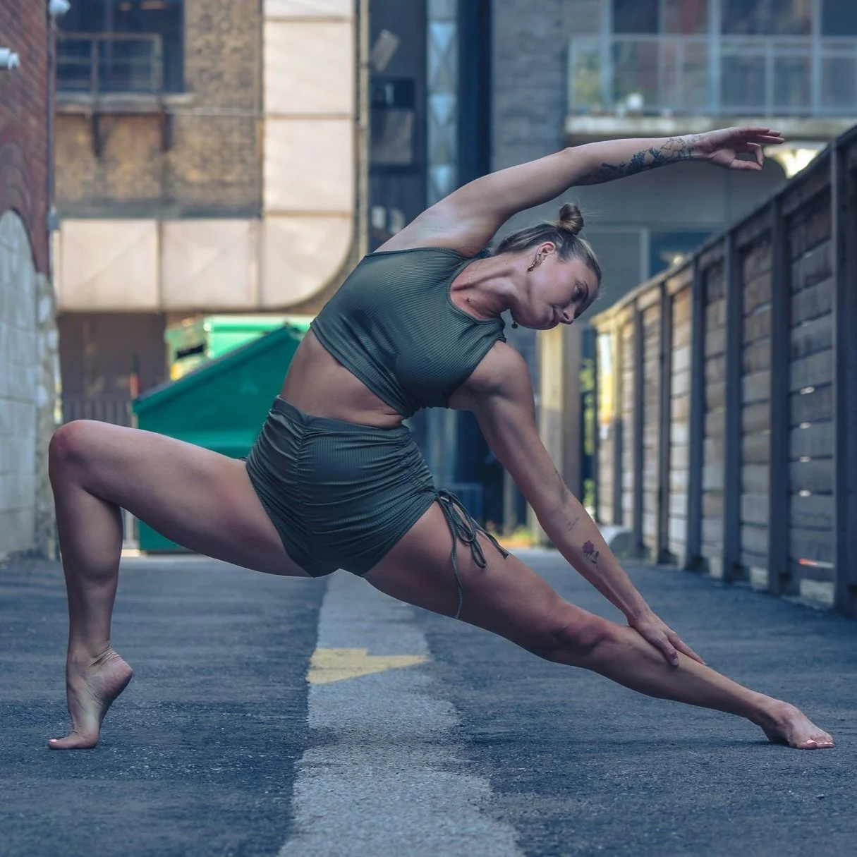 A woman practicing yoga in an outdoor urban alleyway, wearing a dark green athletic outfit, balancing on one foot while extending her arm and leg in a stretch.