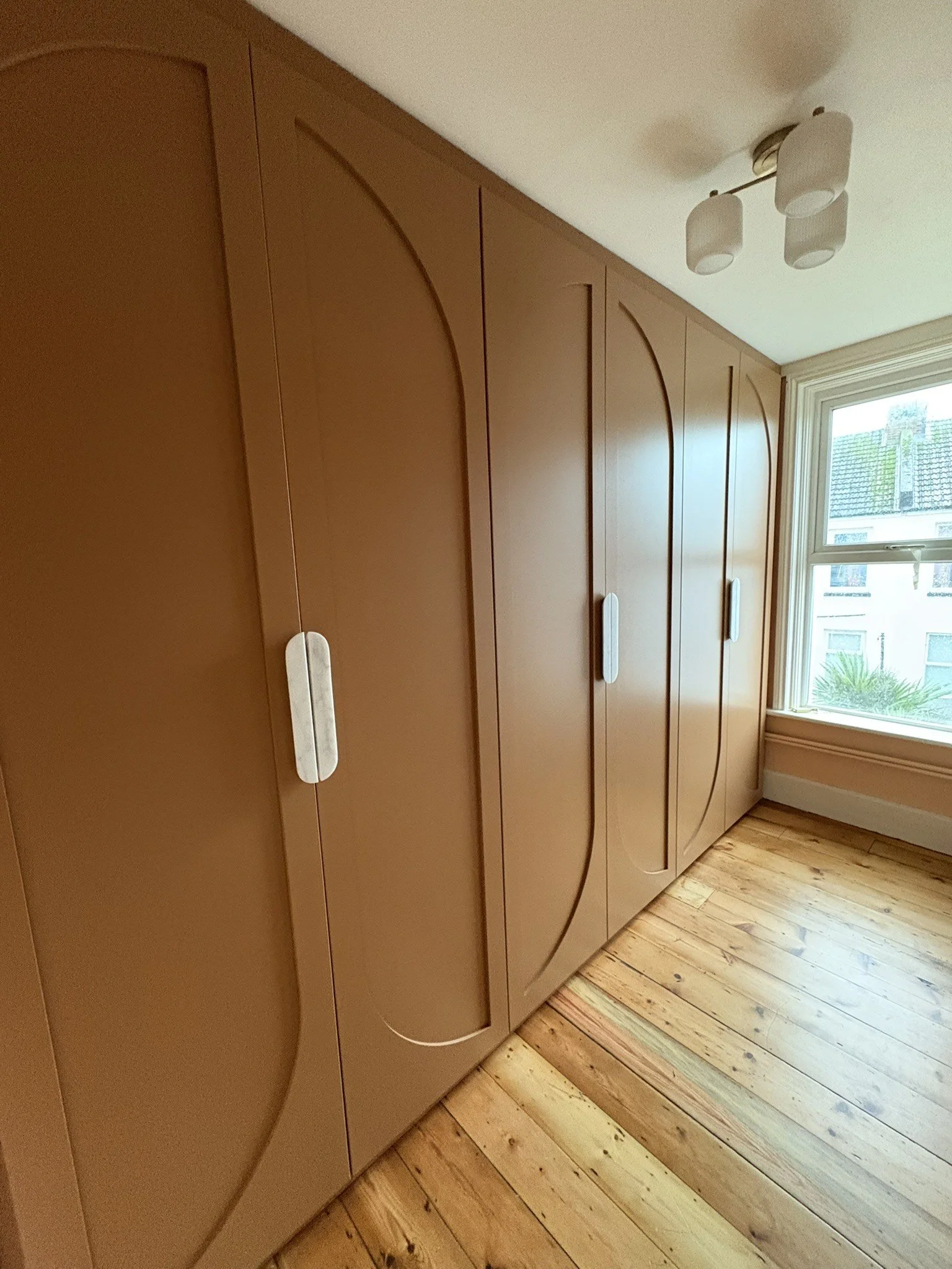 A row of tall wooden cabinets with curved-top doors and silver handles, next to a window in a room with wooden floorboards. Outside the window, a house with a tiled roof and greenery is visible.